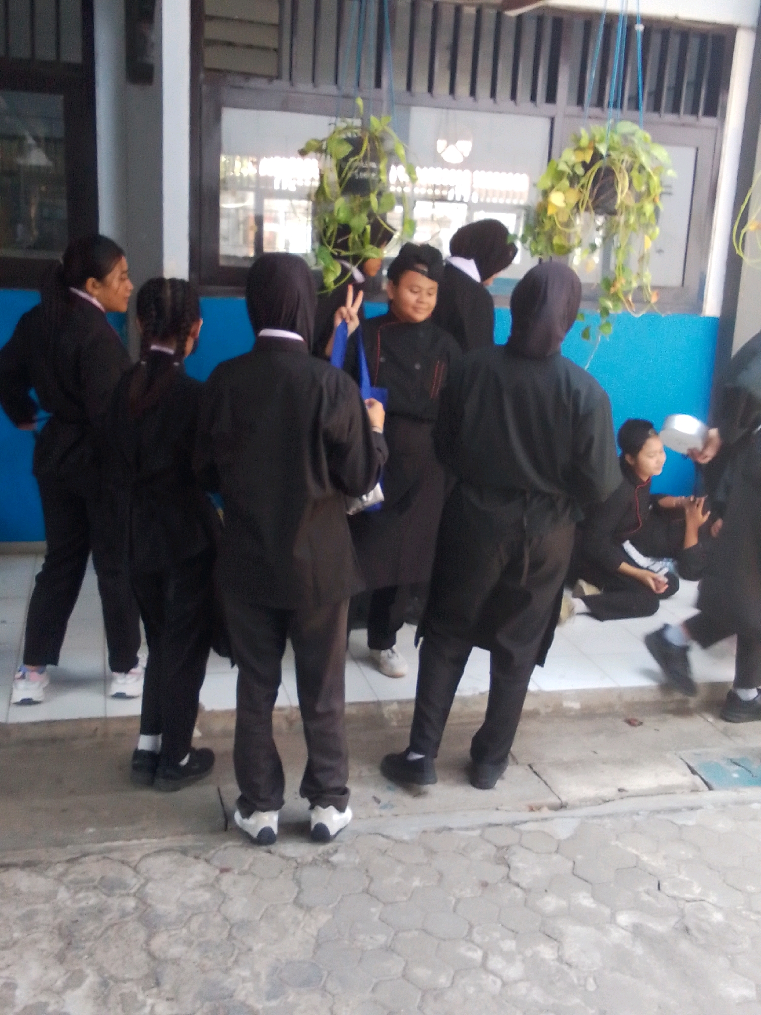 In an outdoor setting resembling a school courtyard or passageway, a group of individuals, predominantly young people in uniform, are gathered. The architecture suggests a building with large windows and overhead structures. Potted plants hang from the ceiling, adding a touch of greenery. The individuals are dressed in dark, possibly school or work uniforms, with some wearing black tops and pants. One person in the foreground wears a head covering. Several individuals appear to be interacting, with some looking towards each other and one holding a bag. The ground is paved with hexagonal tiles. The overall scene conveys a sense of casual gathering or activity.