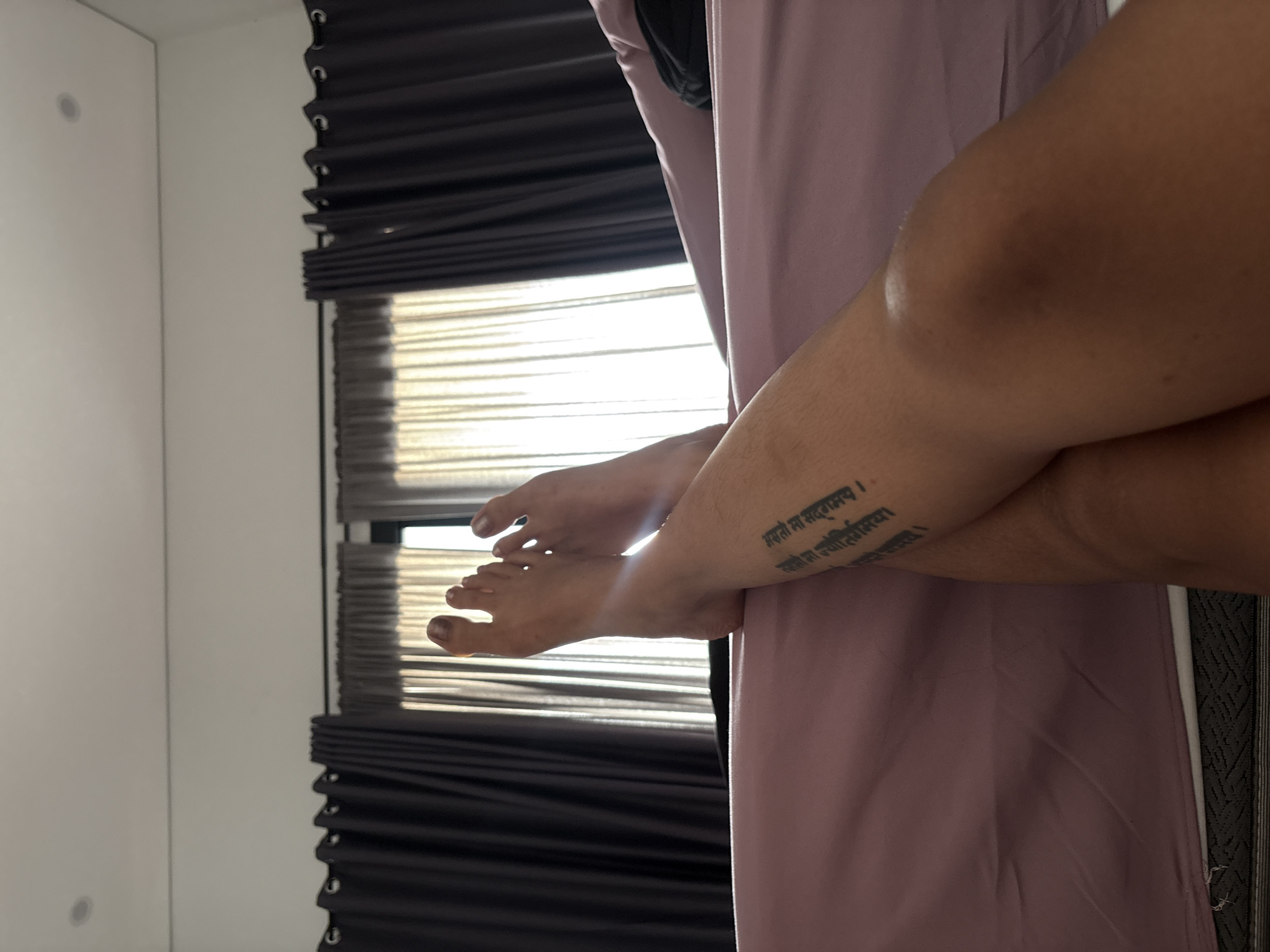 The image displays a close-up, low-angle perspective of a person's legs and bare feet in an indoor setting. The skin tone of the legs and feet is medium, and dark nail polish is visible on the toenails.
A significant detail is a tattoo on the lower right leg, rendered in Devanagari script. The text of the tattoo reads:
"असतो मा सद्गमय ।"
"तमसो मा ज्योतिर्गमय ।"
"मृत्योर्मा अमृतं गमय ।"
This is a Sanskrit mantra that translates to "From untruth, lead me to truth; From darkness, lead me to light; From death, lead me to immortality."
In the background, a window with horizontal blinds is partially visible, allowing bright daylight to illuminate the scene, creating a slight lens flare around the feet. Dark, pleated curtains or blinds are drawn to the sides of the window. A light-colored wall or piece of furniture occupies the left side of the frame. A soft, light purple fabric, possibly clothing or a bedsheet, covers a significant portion of the lower and right side of the image, partly obscuring the upper leg. The overall impression is one of a relaxed posture in a sunlit room during the daytime.