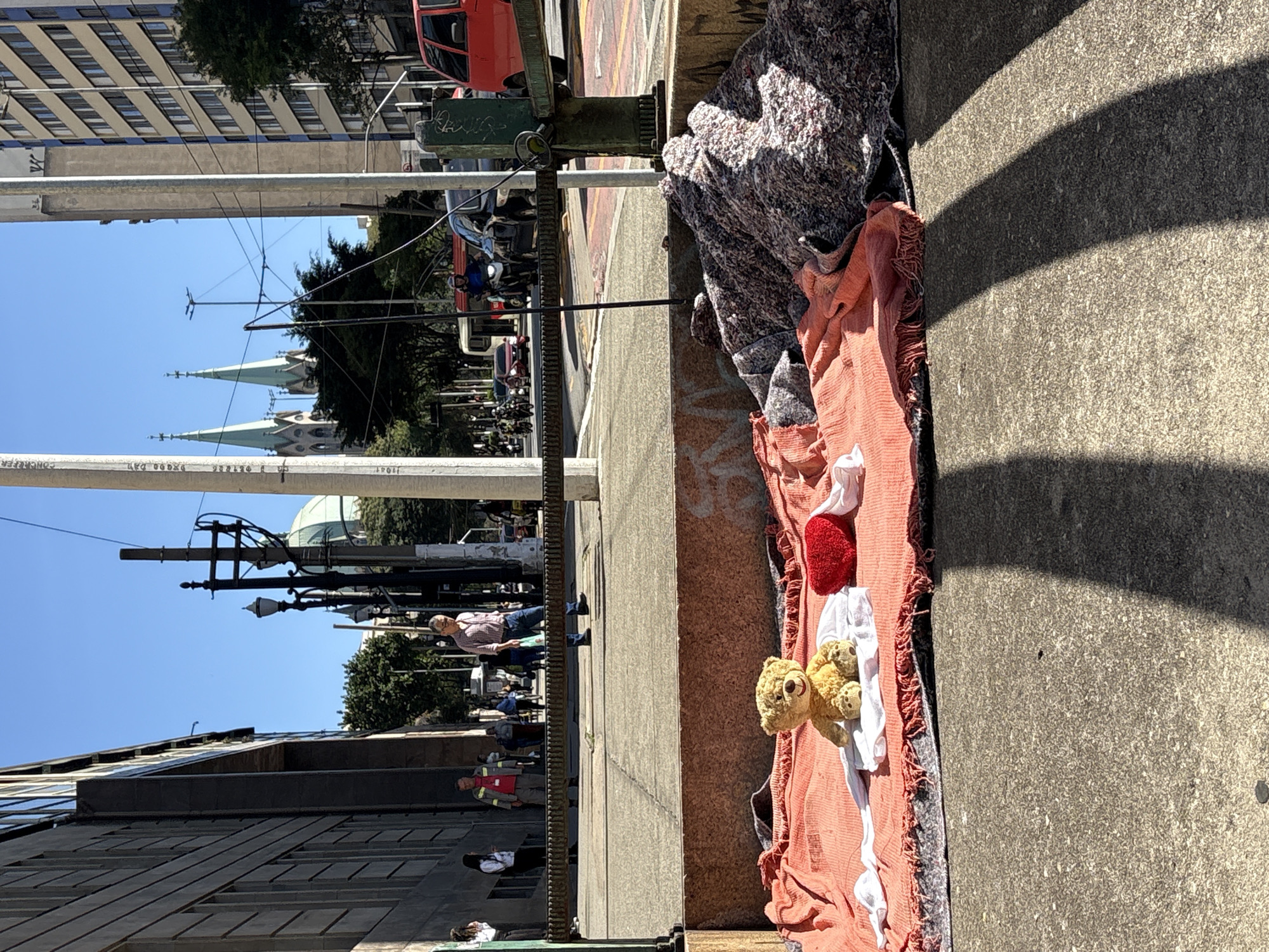 In the foreground of the image, a teddy bear sits on an orange blanket with a white cloth underneath it. To the right of the teddy bear is a red heart-shaped pillow. Behind the blanket are piles of blankets and fabric. The scene is set on a concrete sidewalk in São Paulo, Brazil. In the background, people walk on a sidewalk behind a metal railing. Further back, cars and motorcycles are visible on a street, and a red bus is parked. Tall buildings and trees line the street. In the far background, the distinctive spires of the Cathedral da Sé are visible against a clear blue sky. The image is taken on a sunny day, with shadows cast by an unseen structure.