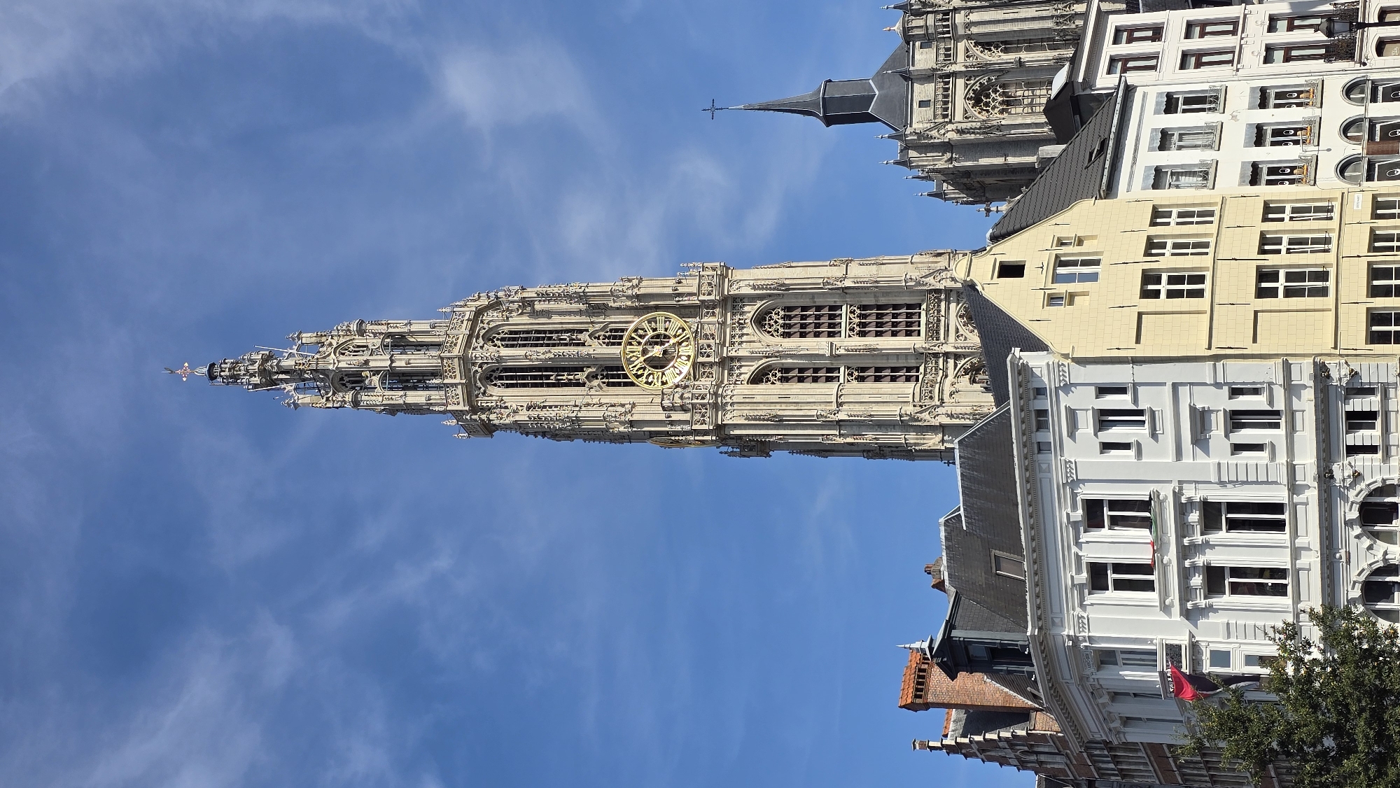 A vertical, low-angle, eye-level outdoor shot captures a towering, ornate Gothic cathedral spire dominating a bright blue sky with wispy clouds. The cathedral's cream-colored stone facade features intricate carvings, tall arched windows, and a prominent, large gold clock face. In the foreground, a row of European-style buildings, with varied architectural designs and pastel colors including white and yellow, are clustered together. These buildings have multiple stories, numerous windows, and pitched roofs, with some showing decorative molding and varied window frames. A small green tree is visible on the left, partially obscuring the lower left corner of the buildings. The scene is one of historical architecture in an urban setting, with the grand cathedral serving as the focal point. The image is located in Antwerp, Belgium, specifically the Cathedral of Our Lady.