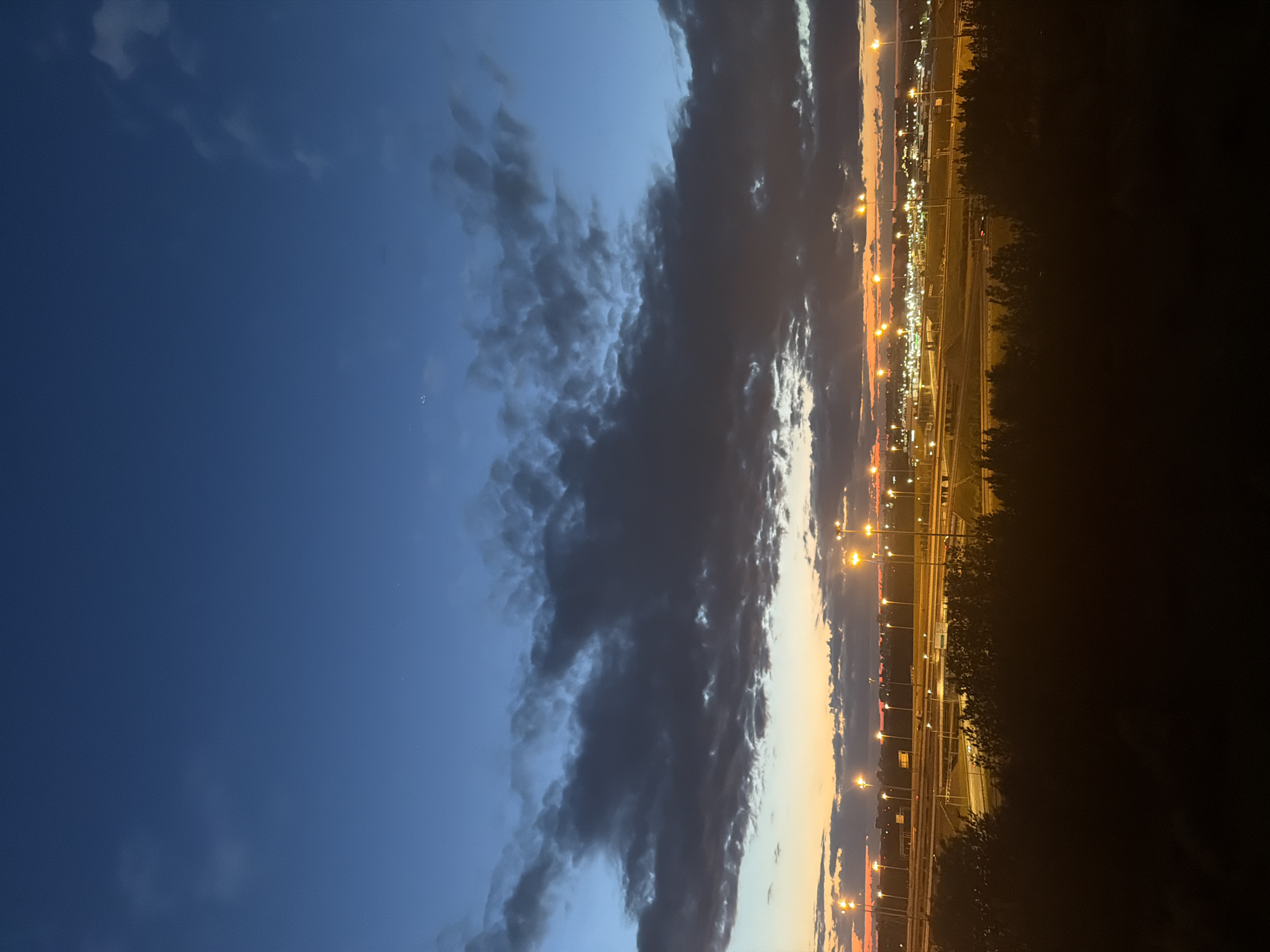 An outdoor, vertical, eye-level shot captures a twilight cityscape. The sky transitions from deep blue at the top to hues of orange and yellow at the horizon, where dark clouds are visible. A line of tall, illuminated lampposts stretches across the mid-ground, casting a warm glow over an urban area. In the foreground, silhouetted trees frame the scene. The location is described as Unknown, Unknown.