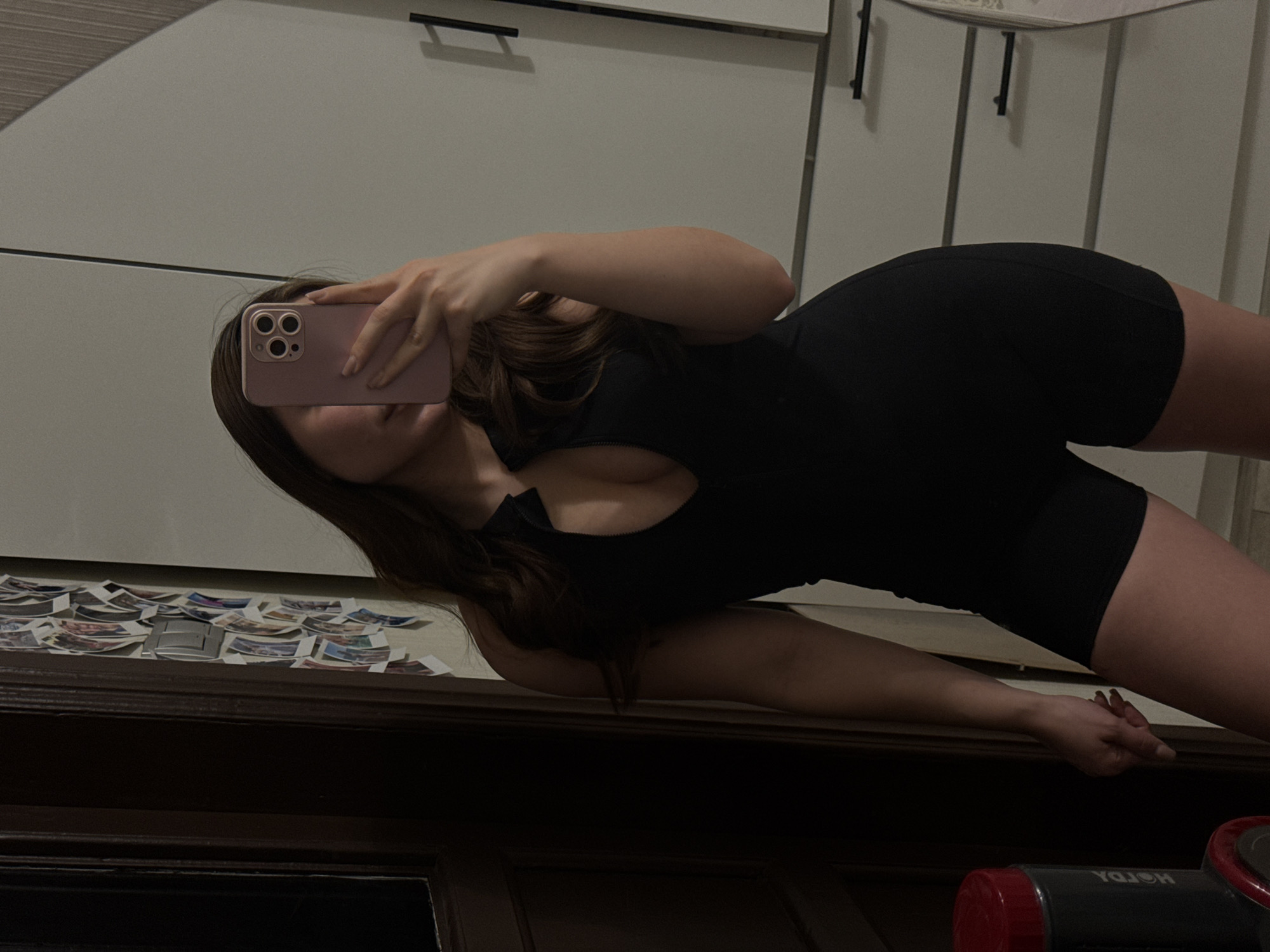 This image is a mirror selfie featuring a woman in an indoor setting, presumably in Kazan, Russia. The woman's face is largely obscured by the pink smartphone she holds, which has a triple-camera system visible on its back. She wears a black, form-fitting outfit, possibly a bodysuit or a combination of a top and shorts, and her long, dark hair frames her face and shoulders.
The scene is reflected in a mirror above a dark wooden piece of furniture, which appears to be a dresser or cabinet. On the top surface of this furniture, numerous small, printed photographs are scattered. The background consists of light-colored (white or light grey) cabinets or wardrobe doors, equipped with simple black horizontal handles.
The image is rotated counter-clockwise, showing the woman in an angled pose. The lighting suggests an indoor environment with artificial light, making the precise time of day unclear, though it could be evening or night. In the bottom right corner, a black bottle with a red cap is partially visible, with reflected text that appears as "YAZON" or similar backwards. No specific emotions are discernible as her face is covered by the phone.