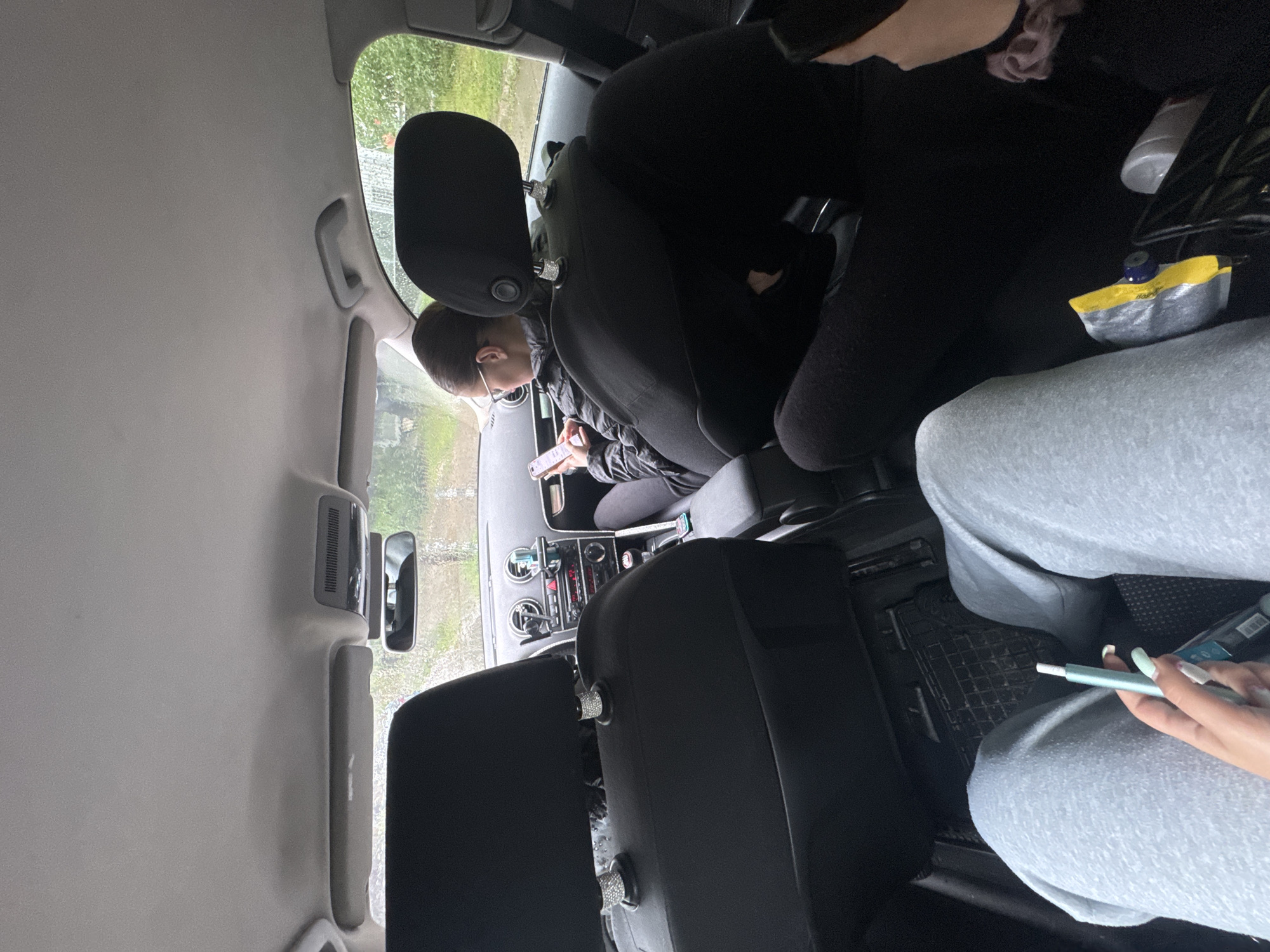 The image, taken inside a car, shows the vehicle's interior from a rear passenger's perspective, looking towards the front. The scene is set in Sofia, Bulgaria, during the daytime, with rain visible on the exterior windows.
Three individuals are present. In the driver's seat, a person wearing a dark jacket and glasses is engrossed in their smartphone. The front passenger is largely obscured, but part of a dark-clothed figure is visible, alongside a water bottle and a yellow-capped pouch with the partial text "HOOP" on the seat next to them. In the foreground, belonging to the back-seat passenger (from whose perspective the photo is taken), legs clad in light grey sweatpants are visible. This individual is holding a light blue vaping device, with an "IQOS" branded pack resting beside it.
Notable details of the car's interior include black headrests, some featuring decorative, possibly diamante, accents on their posts. A portion of the dashboard, revealing controls and vents, can also be seen. The exterior view through the windows shows abundant green foliage, suggesting a natural or suburban environment. The overall activity implies either waiting or traveling in the car, with occupants engaged in personal activities.
