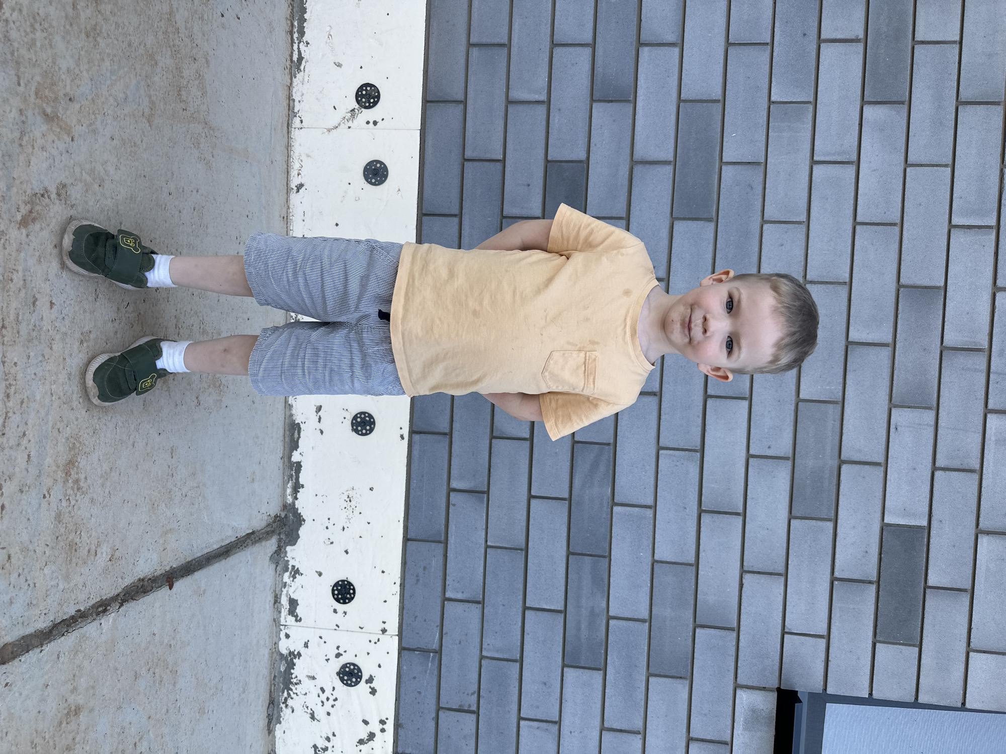 A young boy with blond hair stands on a grey brick surface, wearing a yellow t-shirt, striped shorts, and green shoes. The boy's arms are crossed in front of him, and he is looking directly at the camera. To his left is a textured white surface with black circles, bordering a concrete area. The scene appears to be outdoors. There is no specific evidence to pinpoint the location to the city of Прикамский, Russia.