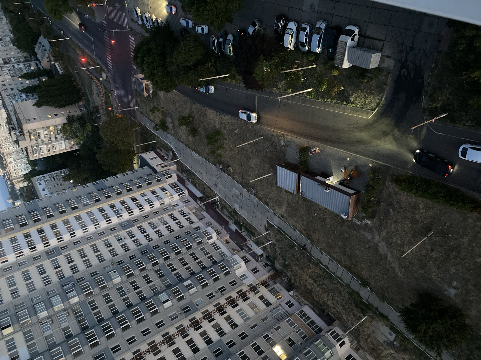 This is an aerial, slightly tilted, high-angle shot of a residential area in Chornomorsk, Ukraine, captured at dusk or twilight. The scene is dominated by a large, multi-story apartment building on the left, characterized by its gray facade and numerous windows. A road runs diagonally through the right side of the frame, lined with parked cars and trees. A pedestrian crossing is visible with red traffic lights illuminated. Two individuals are walking on a paved pathway near the apartment building. Further down the road, several cars are parked in designated spots. The environment appears to be a typical urban setting with a mix of buildings, roads, and greenery.