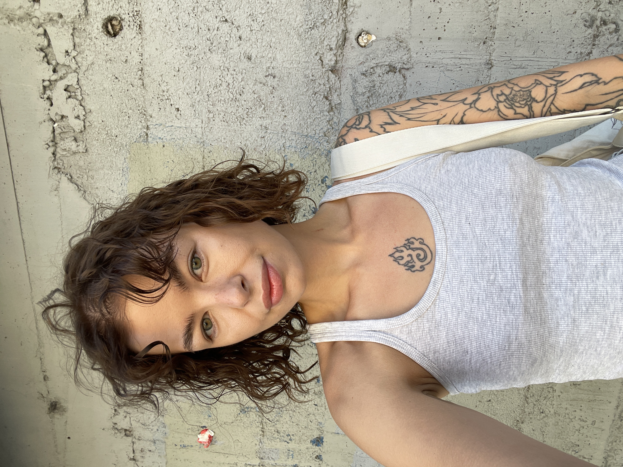 In Rafailovići, Montenegro, a young woman with curly brown hair and green eyes poses in front of a rough concrete wall. She is wearing a light grey ribbed tank top. A tattoo of swirling flames is visible on her chest, and a large floral tattoo covers her left arm. A white canvas bag strap is slung over her right shoulder. The wall behind her is textured and appears weathered, with some smudges and small pieces of debris adhered to its surface. The lighting suggests it is daytime, with sunlight casting subtle shadows.