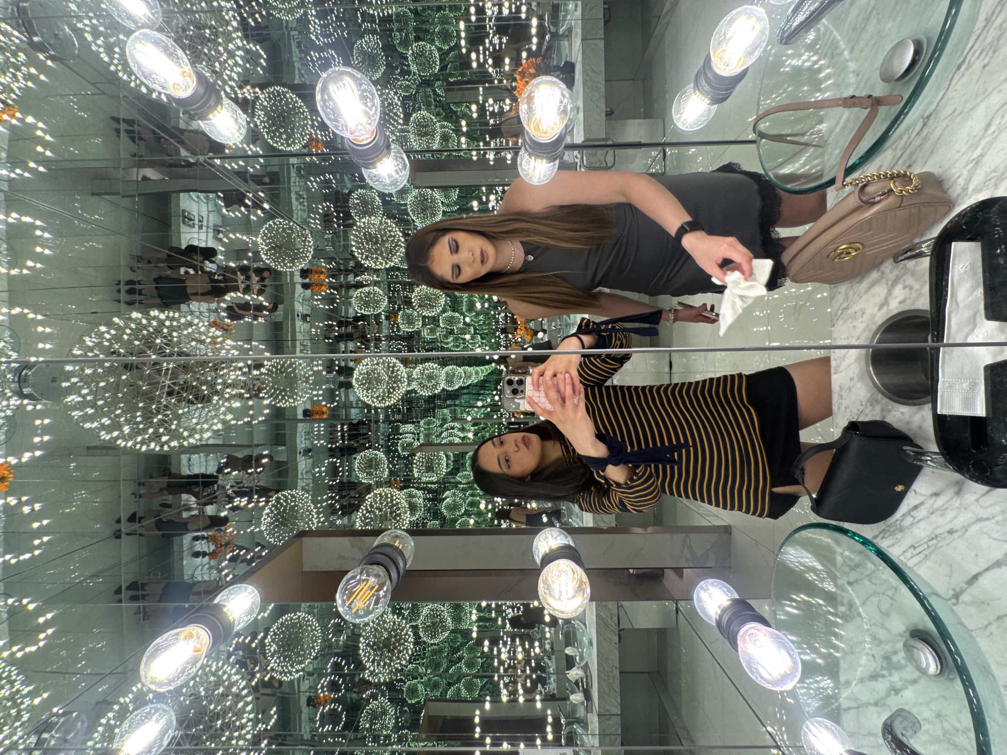 The image appears to be taken in a restroom or vanity area featuring a mirrored ceiling that creates a visual effect of infinite reflections. Two women are visible; one wearing a dark gray dress and holding a white tissue, possibly cleaning a surface. The other is wearing a striped top with dark-colored bows on the sleeves and holding a smartphone, presumably taking a photo.
The surroundings include:
* Several hanging light bulbs and decorative light fixtures, which are reflected in the ceiling.
* Circular glass tables or sinks with marble countertops.
* Personal belongings such as a beige handbag, a black clutch, and a wine bottle.
The environment suggests an upscale establishment. The image might be located in Puebla, Mexico, assuming the user's intent.