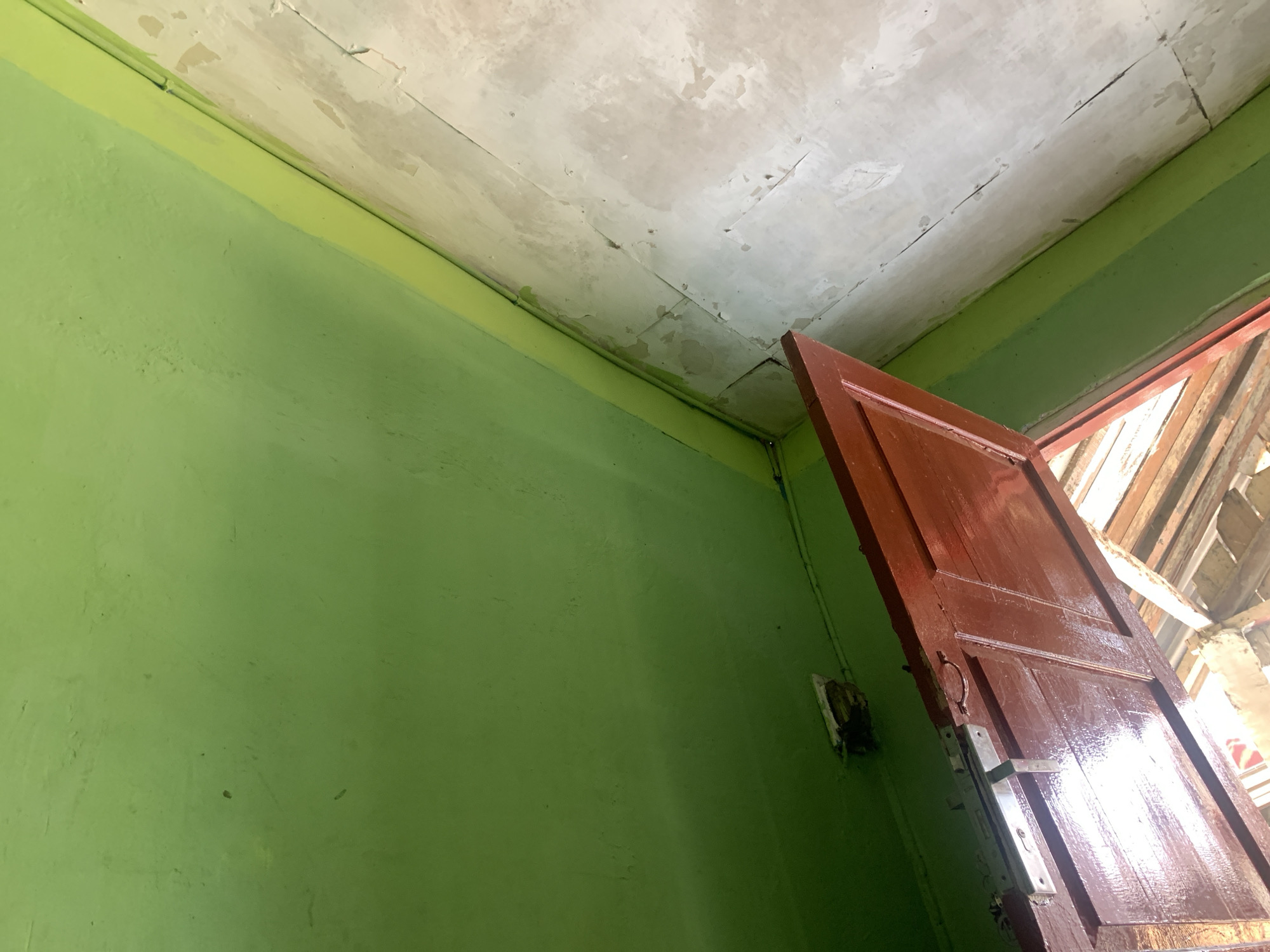 The image shows an interior corner of a room with bright green walls. The ceiling above is white and appears to be made of plaster or a similar material, showing signs of cracking and peeling paint. A reddish-brown wooden door is partially open, revealing wooden beams and what appears to be a section of a roof or attic space beyond. A metal door handle and lock mechanism are visible on the door. A small, square electrical box with visible wiring is mounted on the green wall near the door frame. The scene is lit by natural light coming from the open doorway and possibly from above, casting some shadows. The image was taken in Palembang, Indonesia.