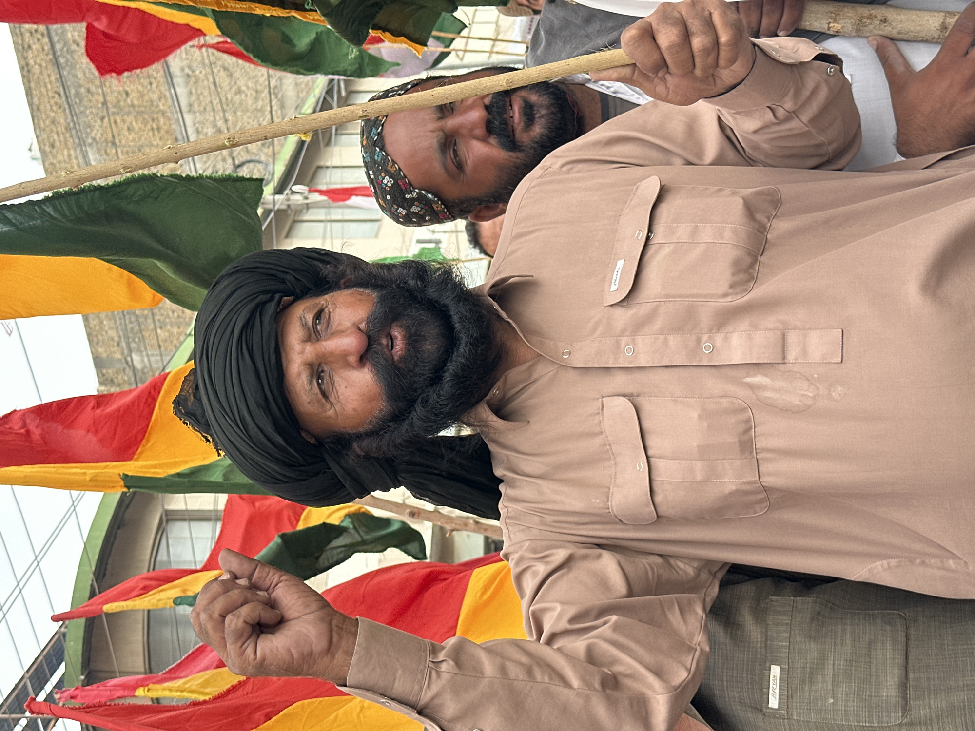 In Quetta City Tehsil, Pakistan, a man wearing a black turban and a tan shirt is shown raising his left fist, holding a wooden stick in his right hand. He has a prominent beard and is looking to his right. Behind him, other men are visible, some holding flags in red, yellow, and green. The environment appears to be outdoors, with buildings visible in the background. The image captures a moment of gathering or protest.