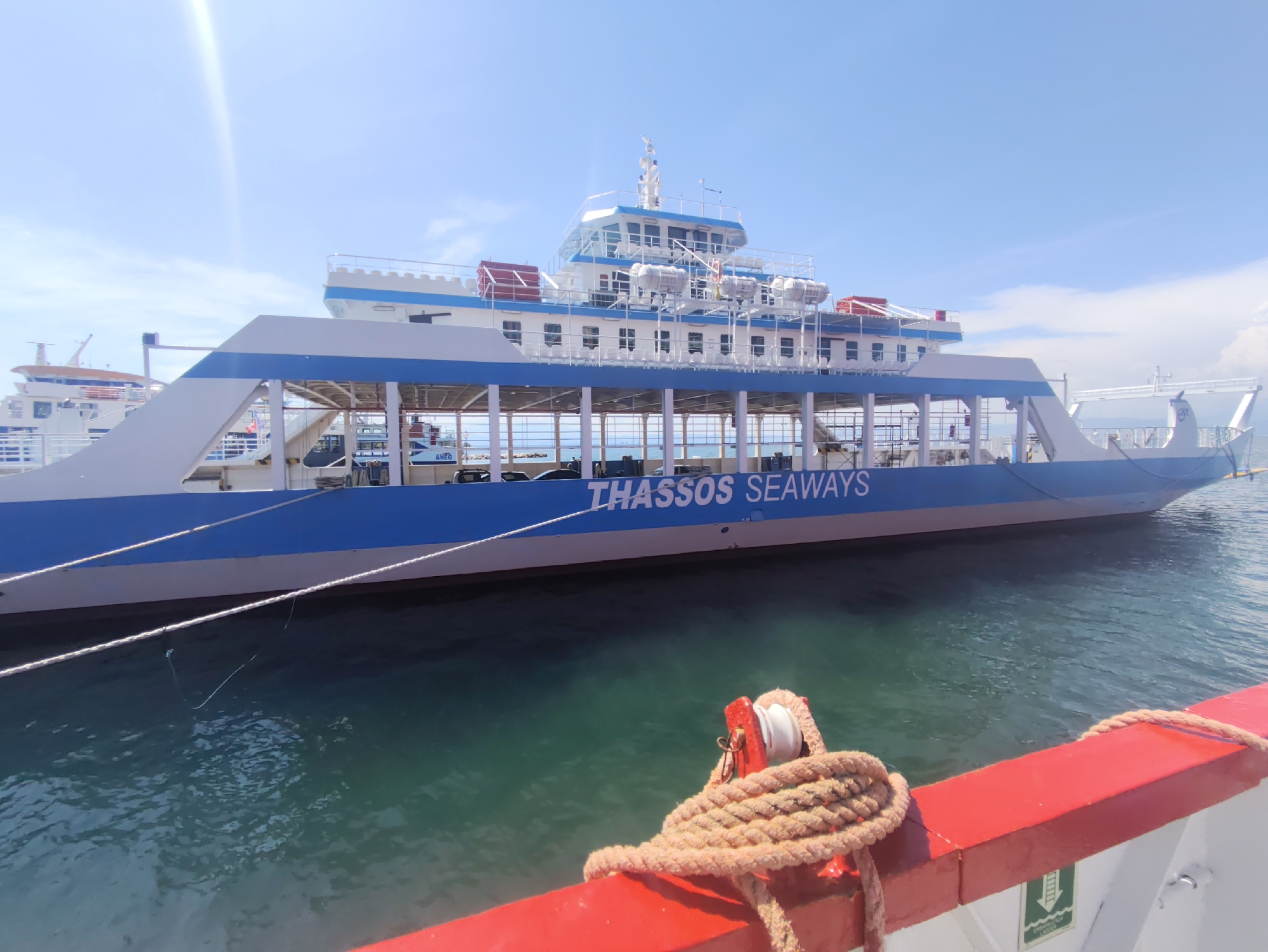 This is a full-frame, eye-level, wide shot of a ferry docked in clear, turquoise water under a bright blue sky with scattered white clouds. The ferry, named "THASSOS SEAWAYS" in blue lettering on its hull, is predominantly white and blue, with a multi-level structure featuring rows of windows. Life preservers are visible along the upper deck. In the foreground, a thick rope is tied to a red railing, indicating the perspective is from another vessel. The water is calm, with slight ripples, and the sun is shining brightly, creating lens flare in the upper left corner of the image. Other ships are visible in the background, suggesting a busy port.