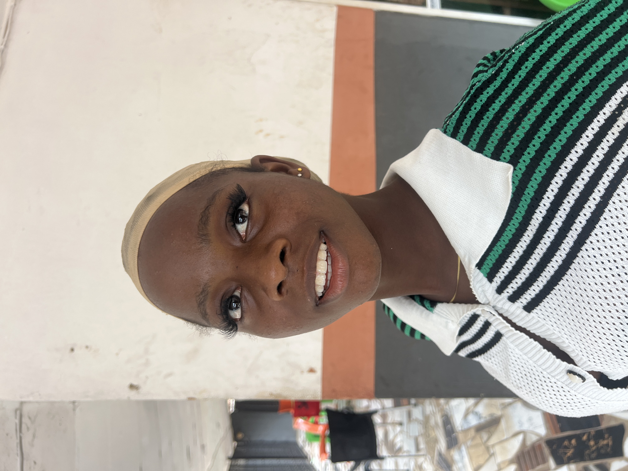 A close-up, eye-level shot features a young Black woman wearing a beige wig cap and a white, green, and black striped polo shirt. She is looking slightly upwards and to her right, with a subtle smile showing her teeth. Her makeup includes long, dramatic eyelashes and small stud earrings. The background consists of a building with textured walls in shades of white, orange, and gray. To the left, a blurred outdoor area shows some chairs and tables, suggesting a public space like a cafe or seating area. The overall lighting is natural and slightly diffused, indicating an outdoor or well-lit indoor setting in Nkpor, Nigeria.