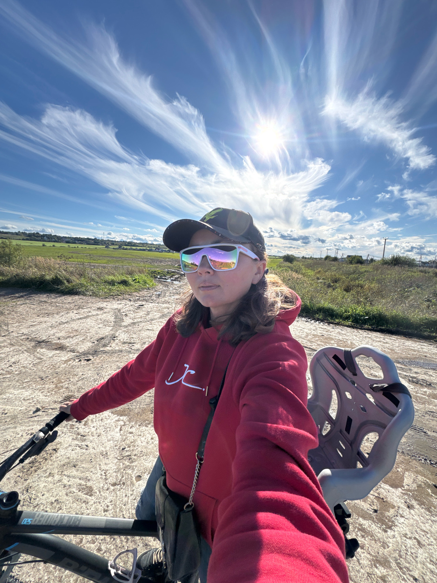 A woman on a bicycle is pictured from a first-person perspective, facing forward, with the sky taking up the upper half of the image. She is wearing a red hooded sweatshirt, blue jeans, a dark baseball cap, and large, reflective sunglasses. Her right hand is on the handlebars of the bicycle, and a portion of the bike, including the frame, handlebars, and a child seat mounted behind the seat post, is visible. The background depicts a dirt road or path, a field with dry grass and some green patches, and a distant line of trees and structures, suggesting a rural or semi-rural setting. The sky is bright blue with scattered, wispy white clouds and the sun is prominently visible, casting lens flare. The sun's rays are strongly illuminating the scene. The location is specified as Kempelevo, Russia.