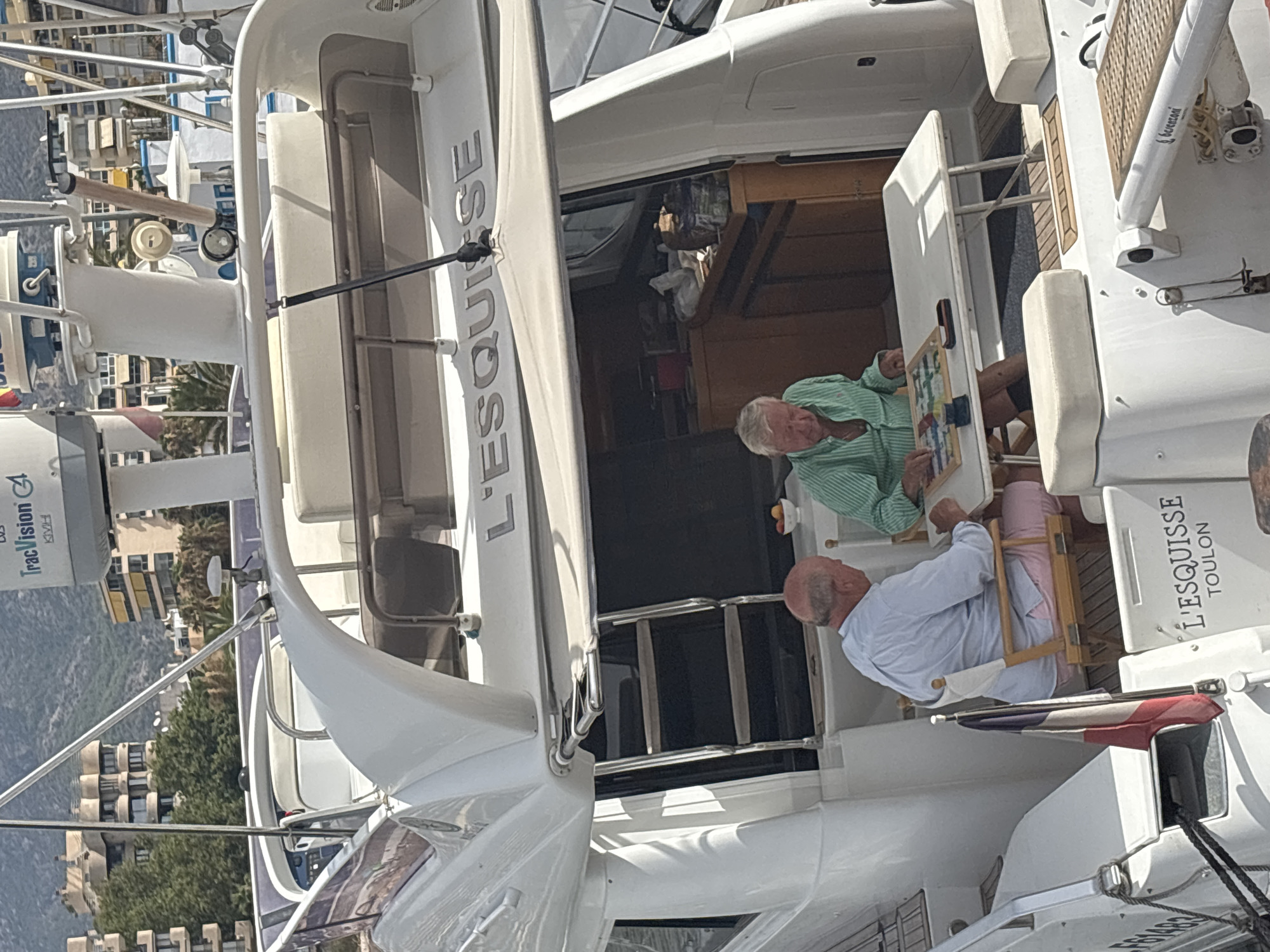 Two elderly Caucasian men are seated at a white folding table on the deck of a white yacht, which is docked in a harbor. The man on the left is wearing a light blue collared shirt and pink shorts. He is seated in a director's chair and looking down at a board game on the table. The man on the right is wearing a green and white striped collared shirt and black shorts. He is also seated in a director's chair, looking at the board game, with his hands on the table. The board game appears to be a type of strategy game with colorful pieces.
The yacht has a white superstructure with the name "L'ESQUISSE TOULON" printed on the stern. A French flag is visible on a small pole near the stern. The background shows buildings and palm trees, suggesting a warm climate. The yacht is surrounded by other boats and structures indicative of a marina. The overall scene is one of leisure and recreation.