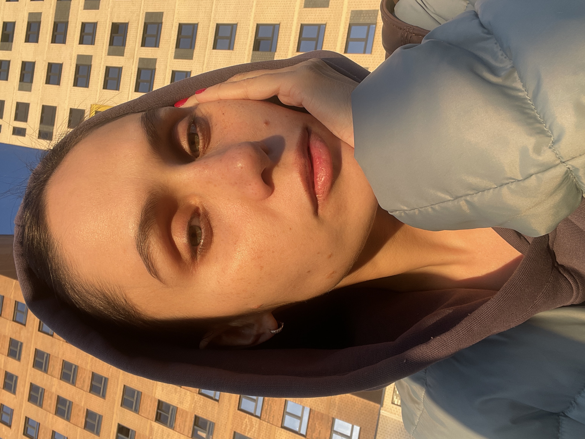 The image is a rotated close-up portrait of a young woman, captured at a 90-degree clockwise angle. She has dark hair mostly covered by a dark grey or brown hooded sweatshirt, which is worn under a light blue puffer jacket. Her face, illuminated by warm, golden sunlight, shows a natural complexion with freckles. Her light brown eyes look directly at the viewer, and her lips are slightly parted. Her right hand, adorned with red nail polish, gently rests near her temple.
The lighting, characterized by strong highlights and shadows, suggests either early morning or late afternoon (golden hour). In the background, a tall, light yellow or tan building with a repetitive pattern of dark, rectangular windows is visible, indicating an urban setting. A clear blue sky is seen in the upper left corner. The subject’s expression is calm and neutral. No text is visible in the image. The scene is consistent with an urban environment, specifically Izhevsk, Russia.