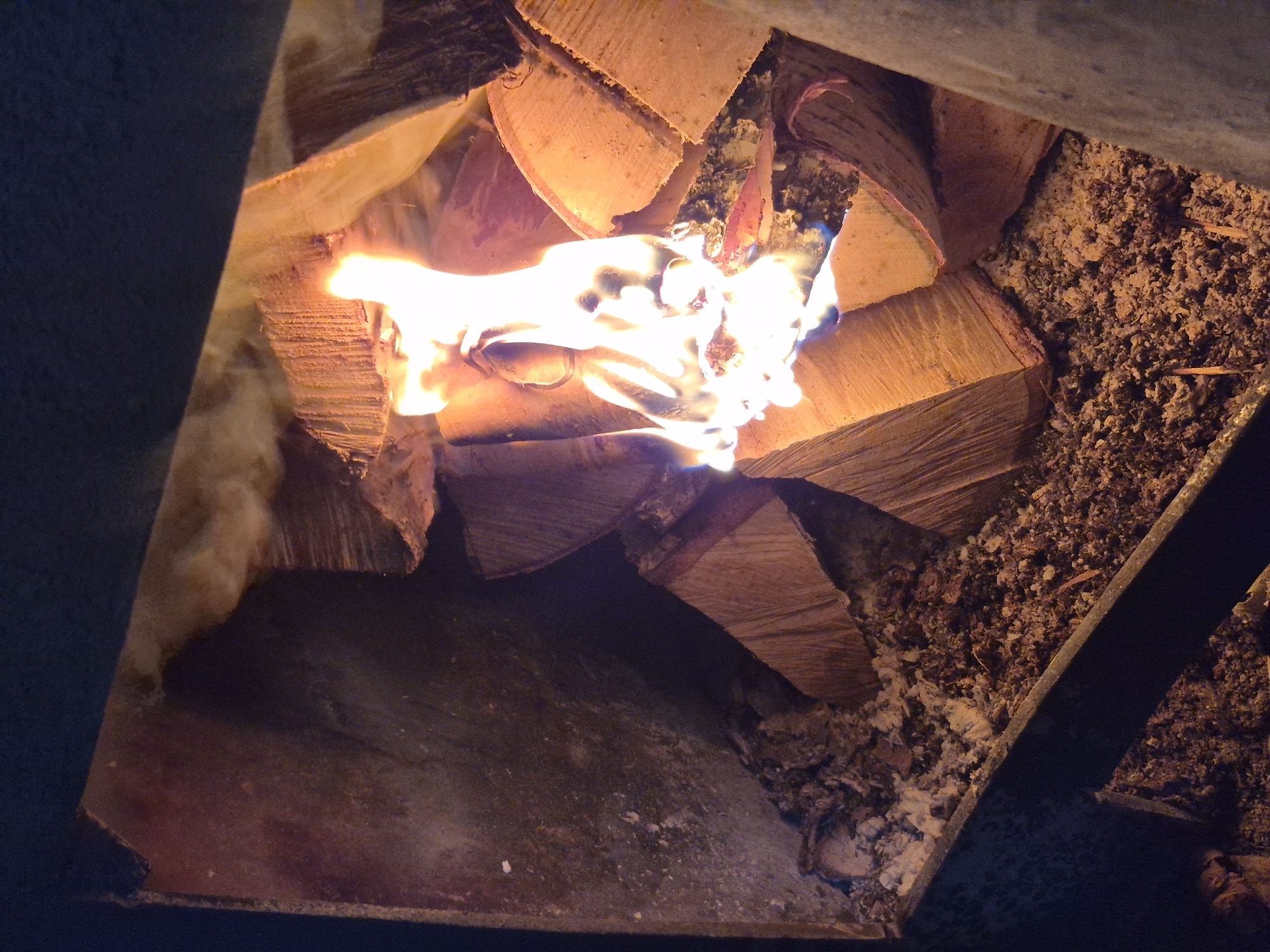 The image shows a close-up view of a fireplace with burning logs. The fire is bright and lively, with flames reaching upwards. Several logs are visible, some stacked and others actively burning. The fireplace appears to be made of metal and is filled with ash and embers at the bottom. The background is dark, suggesting it is nighttime or the fireplace is in a dimly lit room. There is no indication of the city of Kharnavy, Russia, or any people in the image.