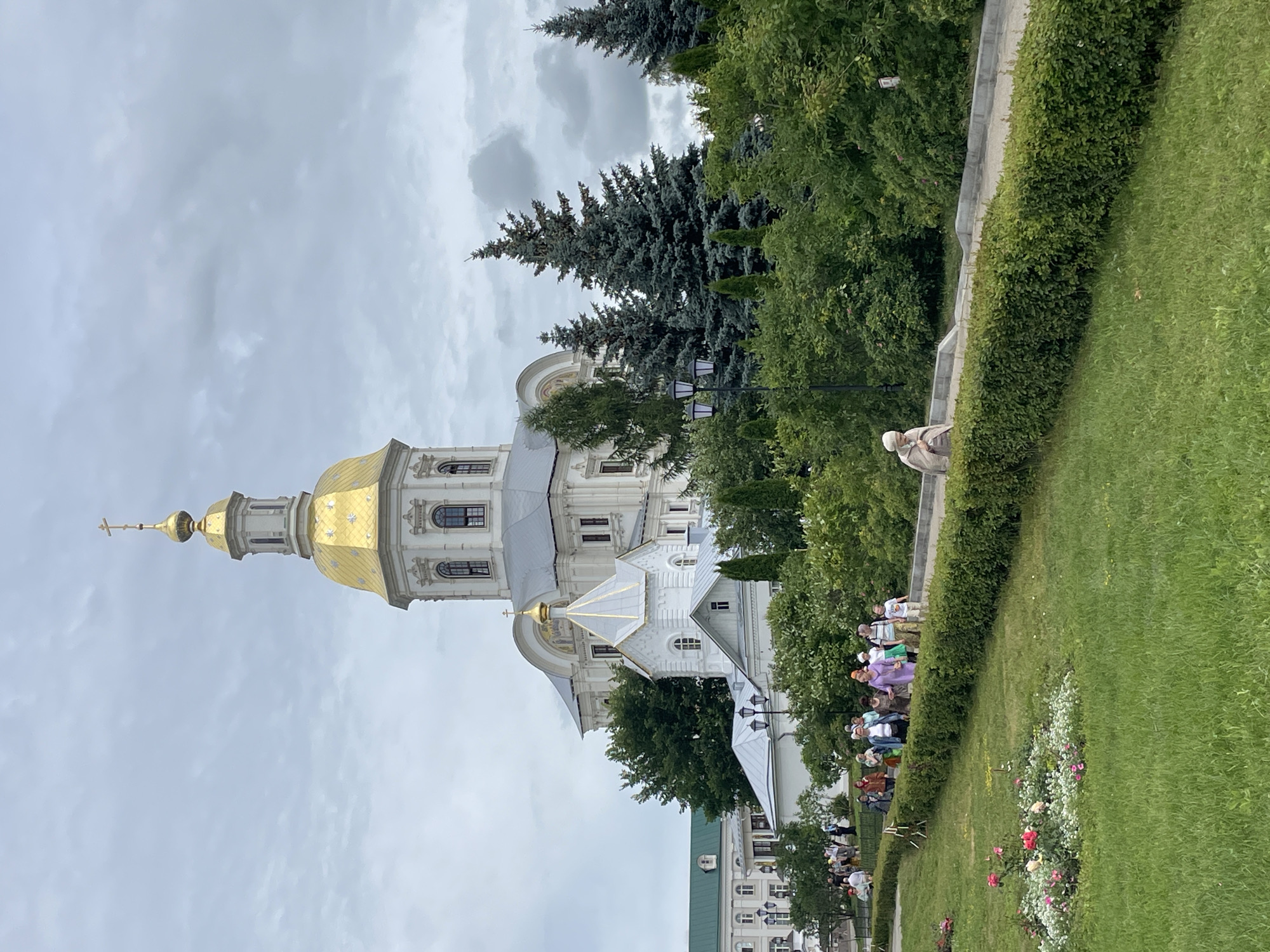 In Diveyevo, Russia, a group of people walk along a paved path bordered by a manicured hedge. In the background, a large white building with a golden dome and cross, likely a church, stands against a cloudy sky. The building features multiple windows and architectural details. Several evergreen trees are visible to the right of the building. In the foreground, a lush green lawn is dotted with a small flower bed containing pink roses and white and pink blossoms. The overall scene depicts a religious or historical site with visitors.