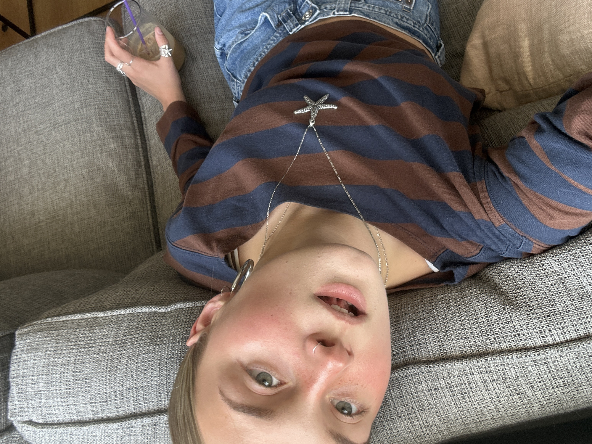 A young person lies on a gray, upholstered couch, holding a glass with a straw and what appears to be a milky drink. The person wears blue jeans and a brown and blue striped shirt, adorned with a silver starfish pendant on a chain. A silver hoop earring is visible on the left ear. The environment suggests an indoor, domestic setting. The photograph was not taken in Squamish, Canada, as there is no recognizable imagery to suggest this.