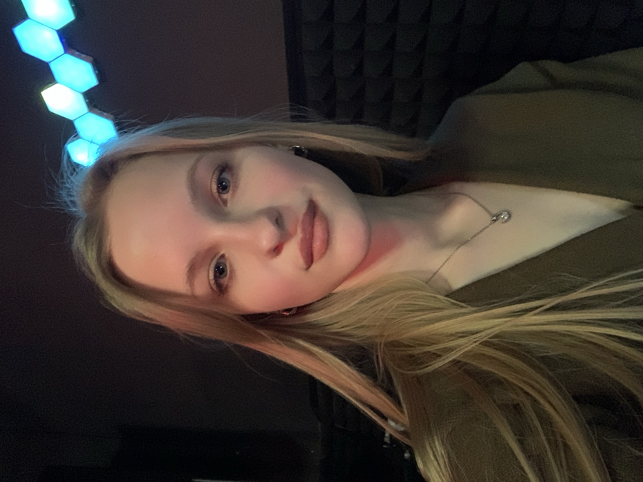 The image shows a young adult female with long, straight blonde hair, fair skin, and blue eyes. She is wearing a dark, possibly olive green or dark brown, V-neck garment and a silver-colored necklace with a small, clear pendant. Her lips appear to have a natural-colored lip product applied. She is positioned centrally, looking directly at the camera. The background is dark. To her right, a dark wall is visible, covered with black acoustic foam panels featuring a pyramidal or wedge pattern. Above and behind her head, a curved arrangement of hexagonal LED light panels emits blue and white light. The lighting in the scene is dim, with light primarily illuminating her face from the front and the blue/white light from the hexagonal panels. No specific visual identifiers within the image directly indicate a location within the city of Kyiv, Ukraine; the environment appears to be an indoor space such as a recording studio or a room with soundproofing.