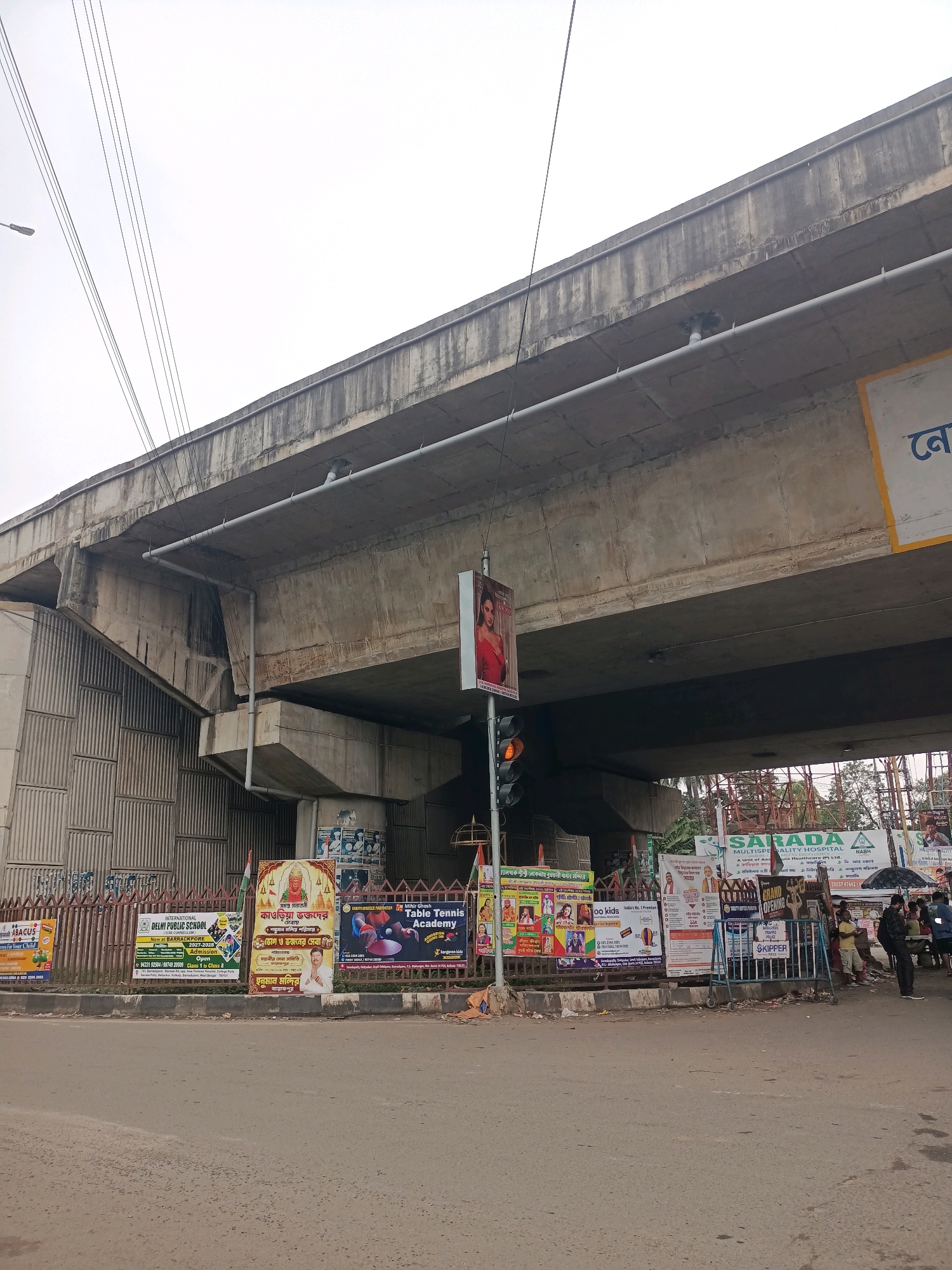 The image shows a street scene in Barrackpore, India, with a flyover dominating the upper portion of the frame. Several posters are displayed along the roadside beneath the flyover. One poster advertises "Mihir Ghosh Table Tennis Academy". Another poster promotes "INTERNATIONAL DELHI PUBLIC SCHOOL" and lists admission details for Barrackpore. A poster for "SARADA MULTISPECIALITY HOSPITAL" is also visible. A traffic signal with orange and red lights is present on the left. Several people are standing or walking near the posters and a black umbrella is held by someone. The environment appears to be an urban street with a road in the foreground and a concrete flyover overhead. The sky is overcast.