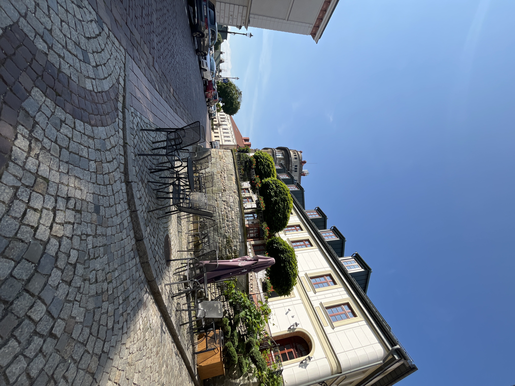 The image depicts a street scene in a European city, possibly in a historic district, given the cobblestone pavement and architecture. The perspective is from street level, looking up a gentle incline. On the right side of the frame is a cream-colored building with a dark tiled roof and dormer windows. This building has red-framed windows and an arched entrance with a red door. In front of this building, there is an outdoor seating area for a cafe or restaurant, with several metal tables and chairs, and a large closed umbrella. The seating area is on a raised platform paved with cobblestones. Behind the seating area, a stone retaining wall supports a garden with manicured trees and flowering plants.
To the left of the scene, the street is lined with parked cars, mostly sedans, on the left side. The street itself is paved with cobblestones that transition from a light gray and tan pattern in the foreground to darker gray further up the street. In the background, more buildings are visible, including a distinctive rounded building with several levels and architectural details. The sky is clear and blue, indicating a sunny day. The overall impression is of a charming and well-maintained urban environment.