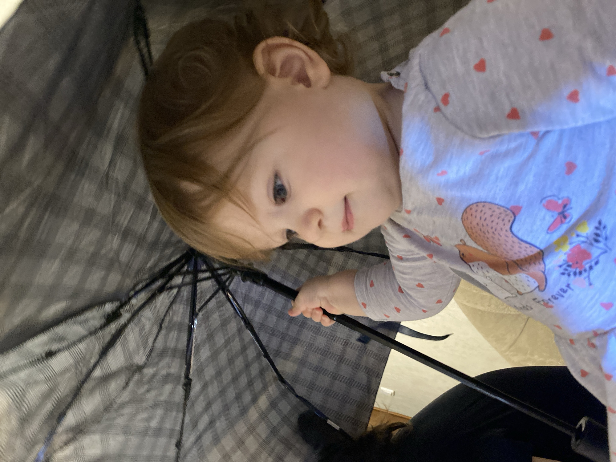 A toddler holds a gray umbrella with a black handle and ribs. The toddler is wearing a white shirt with red hearts and a squirrel and butterfly design. The environment is unknown.