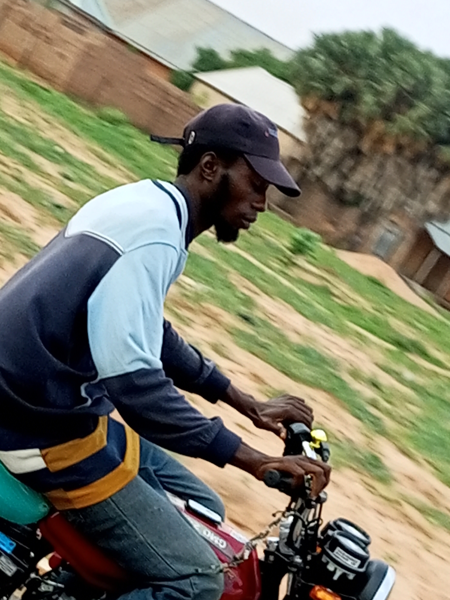 A young Black man wearing a dark baseball cap, a dark blue and light blue long-sleeved shirt with a yellow stripe across the chest, and blue jeans is riding a red motorcycle. He is leaning forward, gripping the handlebars. The background is a blurred landscape of green grass and brown dirt, with a building visible in the distance. The overall scene is captured from a slightly elevated, side angle, suggesting movement. The location is identified as Tempure, Nigeria.