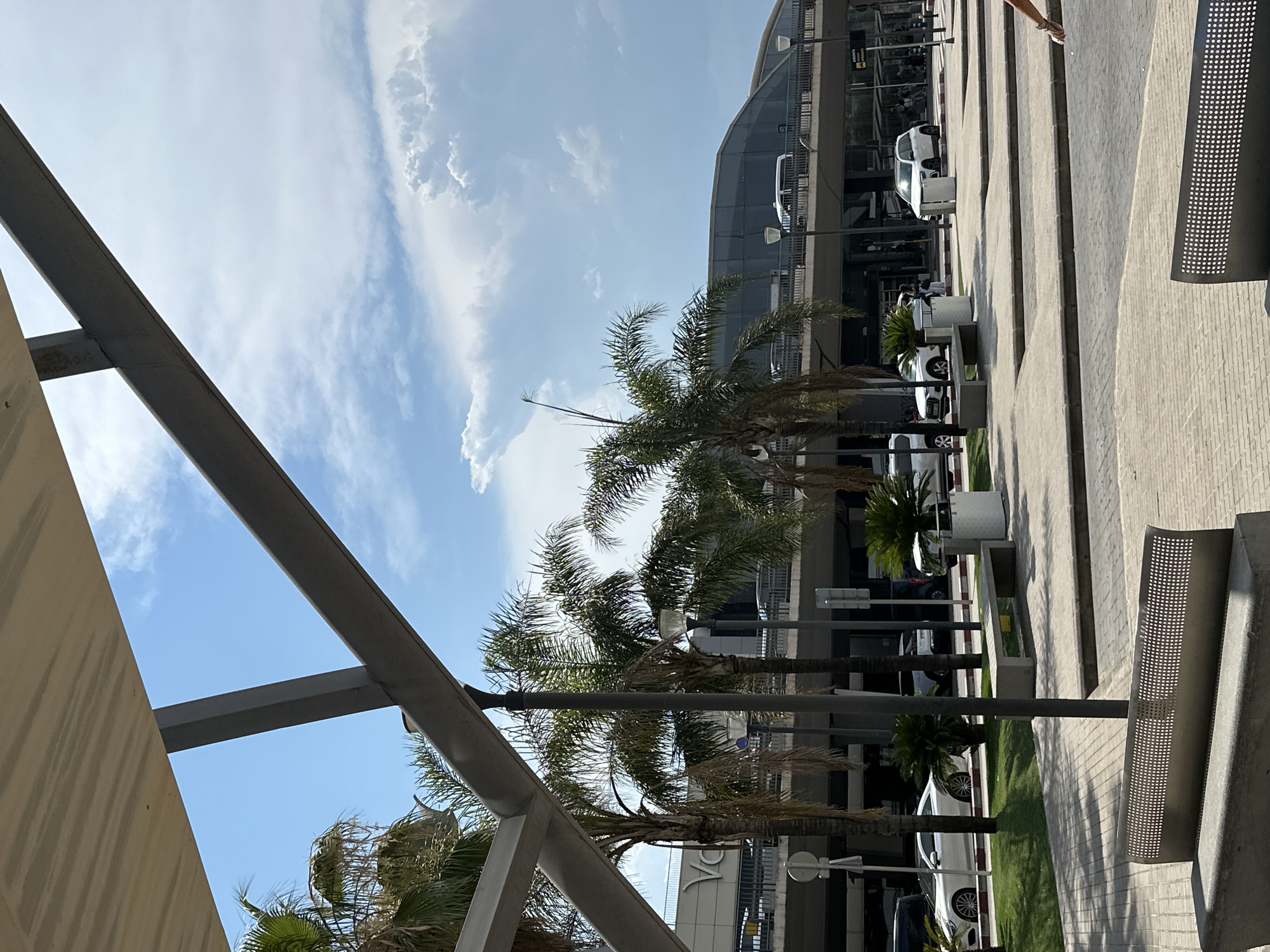 The image shows an outdoor scene in the city of Manises, Spain, likely at an airport due to the modern architectural style of the building and the presence of parked cars. In the foreground, two modern metal benches with perforated seats are visible, placed on a paved area with a brick-like pattern. Behind the benches, a grassy area is seen with palm trees and some smaller potted plants. Several cars, including a white Mercedes-Benz S-Class, a white Audi SUV, and other vehicles, are parked along the edge of the grassy area and on a road in front of a multi-level building.
The building appears to be a terminal with a curved, glass facade on the upper level, suggesting a modern airport or transportation hub. The sky is blue with scattered white clouds, indicating fair weather. People are visible walking in the background near the terminal entrance. The overall impression is one of a bright, sunny day at a well-maintained public space.