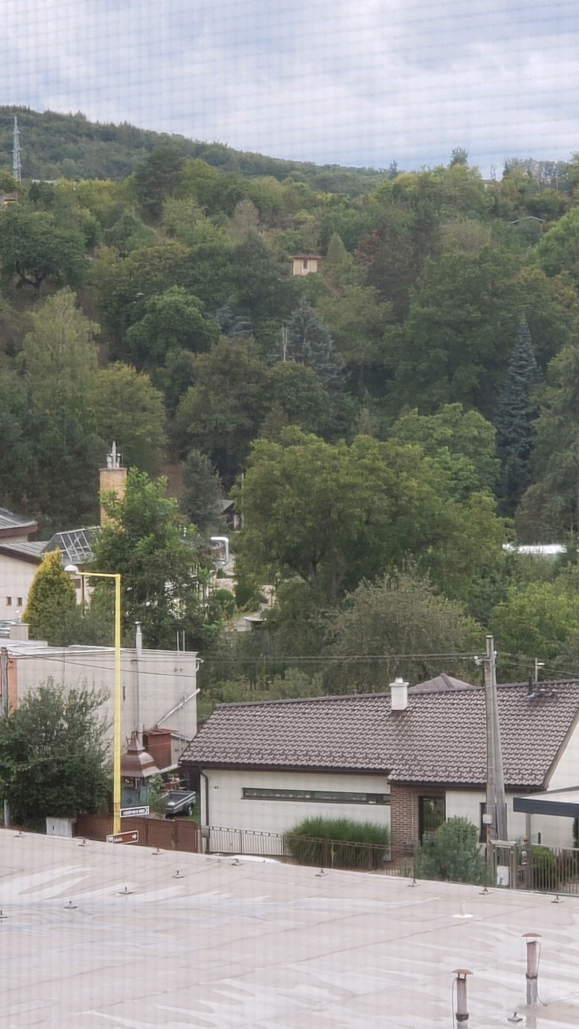 The image is a vertical, eye-level shot taken from an elevated position, possibly a window or balcony, overlooking a residential area nestled against a wooded hillside. In the foreground, a light-colored, flat roof with numerous small, raised fixtures dominates the lower portion of the frame. Beyond this roof, a single-story building with a brown tiled roof and white walls is visible. This building has a long, narrow horizontal window and a smaller, dark-paned entrance. A metal fence encloses a section of the yard, and dense green shrubbery is planted near the building. Further to the left, another light-colored building with a sloped roof is partially visible, with a solar panel array on its roof. A tall, yellow pole stands in front of this building. Behind these structures, the landscape rises into a densely forested hill. Several trees of varying heights and species populate the hillside, interspersed with glimpses of smaller buildings and structures, including a small, light-colored house with a reddish-brown roof nestled amongst the trees. A tall, slender telecommunications tower is visible on the far left of the hillside. The sky is overcast with a light gray hue. The scene appears to be a quiet, suburban or semi-rural setting. Based on the visual cues, this scene is likely located in the city of Prešov, Slovakia, given the architectural style and the surrounding natural environment which is consistent with the foothills of the Carpathian Mountains surrounding Prešov.