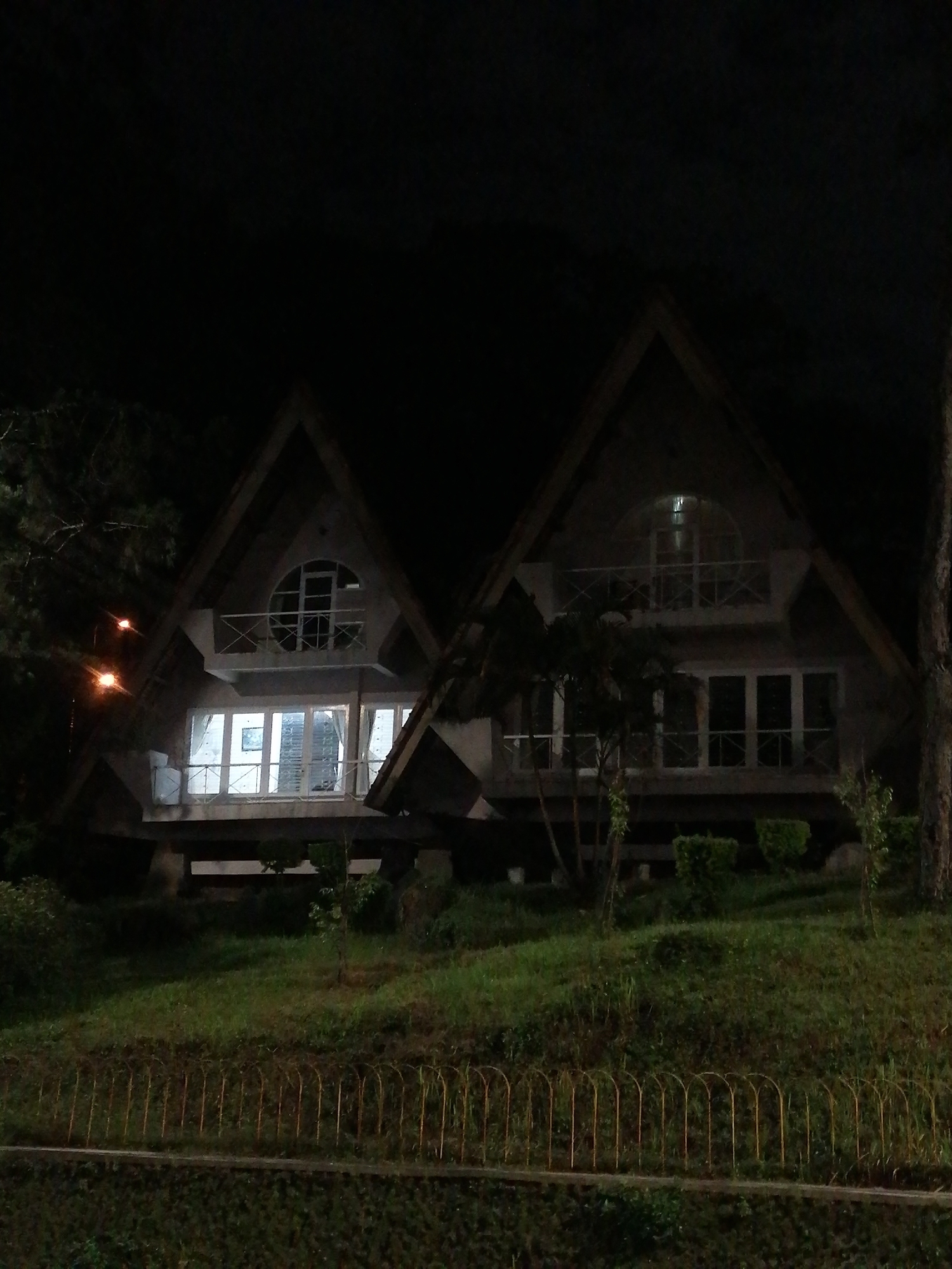 The image depicts two similar A-frame houses at night, likely located in Da Lat, Vietnam. The houses have a triangular roof design and are two stories tall. Both structures feature large windows on both floors, with a rounded window on the top floor of the first building. They have balconies on the second floor with railings. The buildings appear to be built on stilts. The surrounding environment consists of a grassy slope with sparse vegetation, bordered by a metal fence and a stone wall. A few trees are visible, and the sky is dark, indicating nighttime. A streetlight is visible near the first building, casting a warm glow.