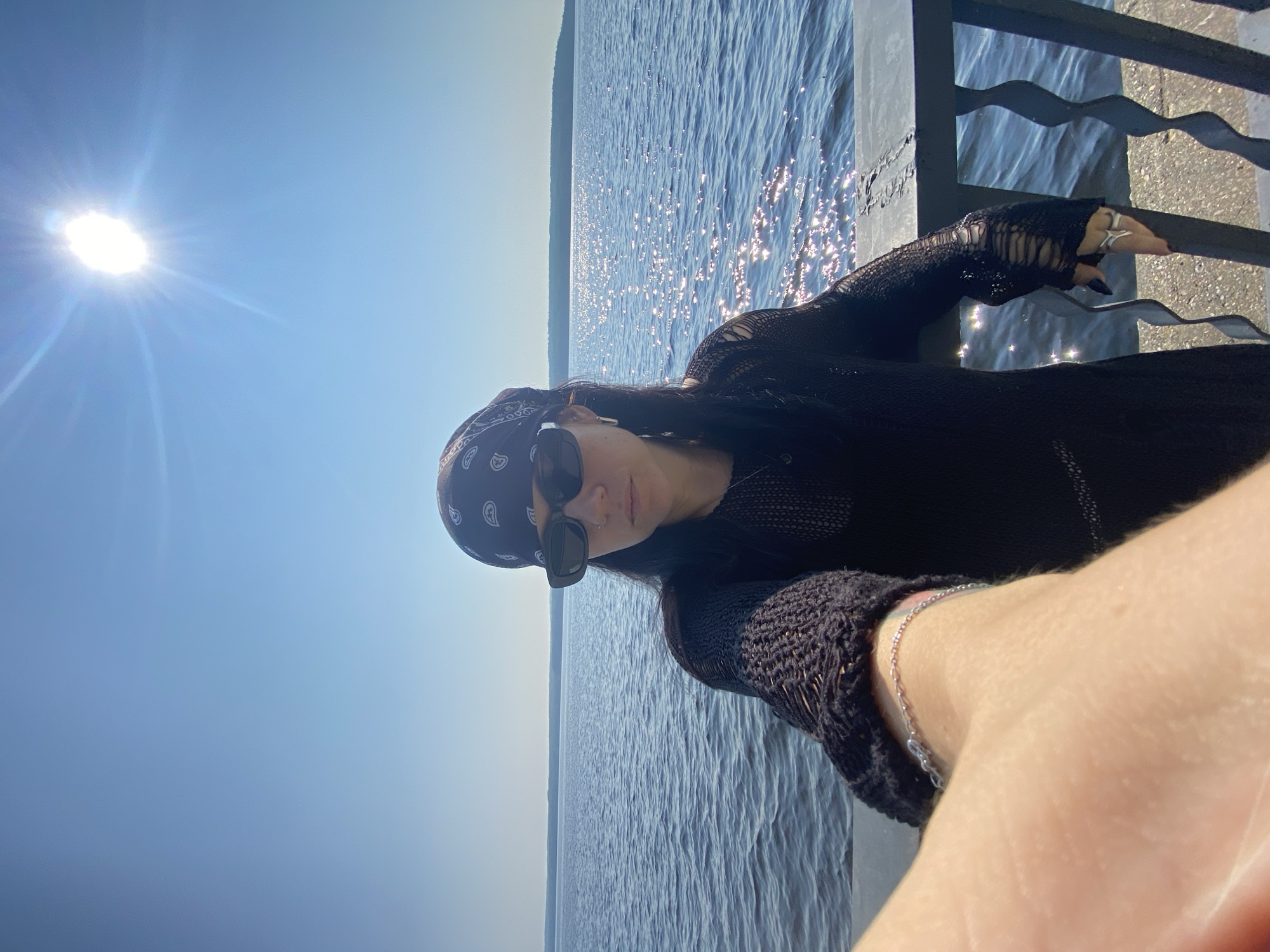 A woman with dark hair, wearing a black bandana and sunglasses, stands on a waterfront promenade. She is taking a selfie, with her left arm extended towards the camera. She is dressed in a black, sheer, long-sleeved top with ripped detailing at the elbows and a silver bracelet on her left wrist. She wears a silver ring on her right index finger. The background features a bright blue sky with the sun prominently visible, casting a glare on the water. A calm body of water stretches to the horizon, where a distant tree line is visible. The promenade is bordered by a dark metal railing. The location is the embankment of the Kama River in Chaikovsky, Russia.