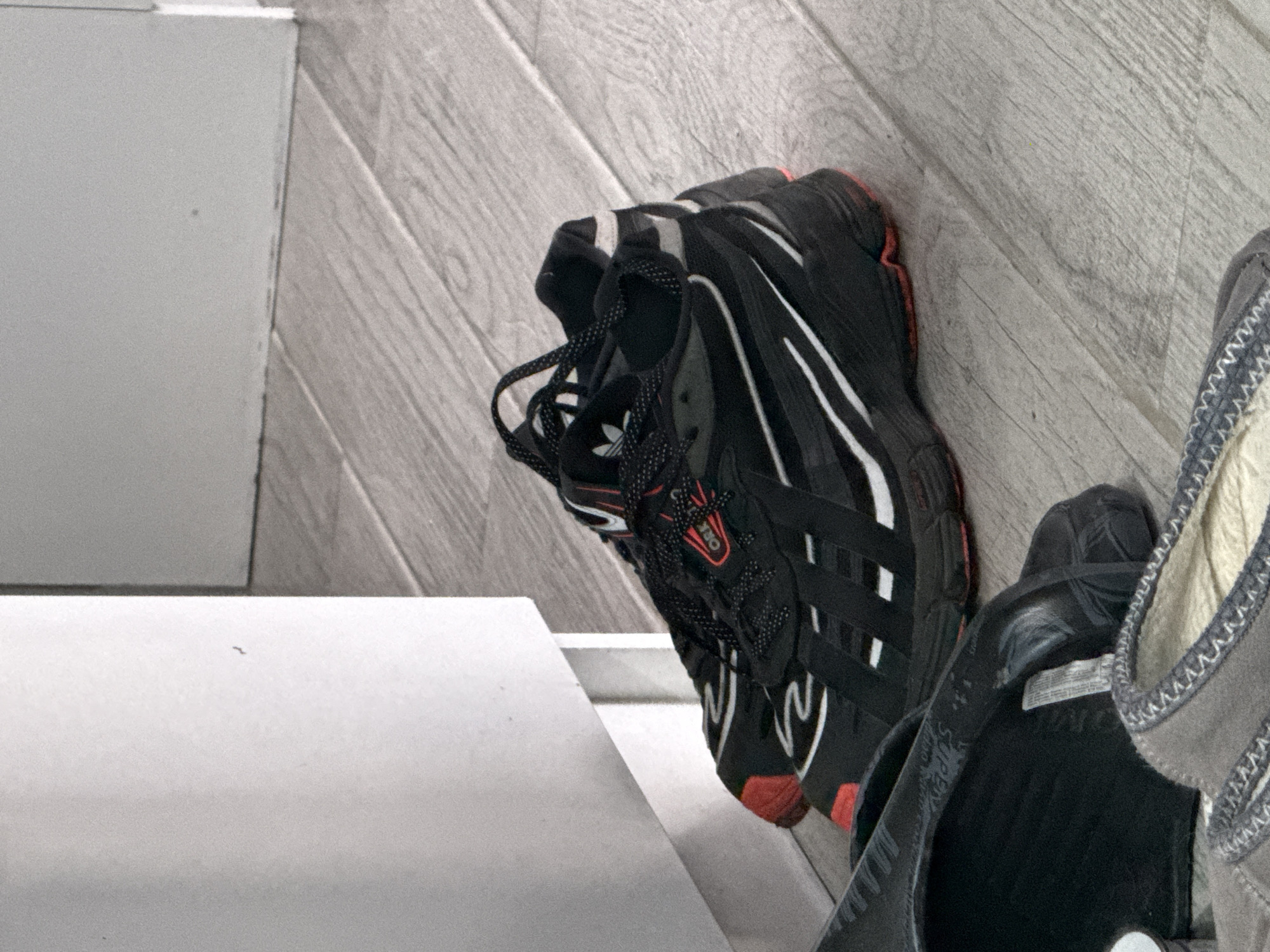An overhead, angled shot captures two pairs of footwear on a light grey wooden floor, placed against a white surface, possibly a wall or baseboard. On the left, a flat, white structure, appearing as a step or low platform, extends into the foreground.
The main subject is a pair of black athletic shoes, likely sneakers, featuring white stripes or accents along the sides and red details on the outsoles and eyelets. The laces are black with small white dots. A small Adidas trefoil logo is visible on the tongue of the left shoe. On a red patch near the laces, faint text, possibly "ORK", can be seen.
To the right, partially visible, is another pair of footwear, appearing as light grey or beige casual shoes or slippers, with a darker grey, textured trim and visible stitching along the edge. A white tag or label inside this footwear contains unreadable text.
The wooden floor, possibly laminate or light hardwood, has planks laid diagonally across the frame. No activity is depicted; the shoes are stationary, suggesting they are stored or temporarily set aside in an indoor environment, likely a home or entry area in Stara Zagora, Bulgaria. The lighting is ambient, indicating an indoor setting without specific time-of-day cues.