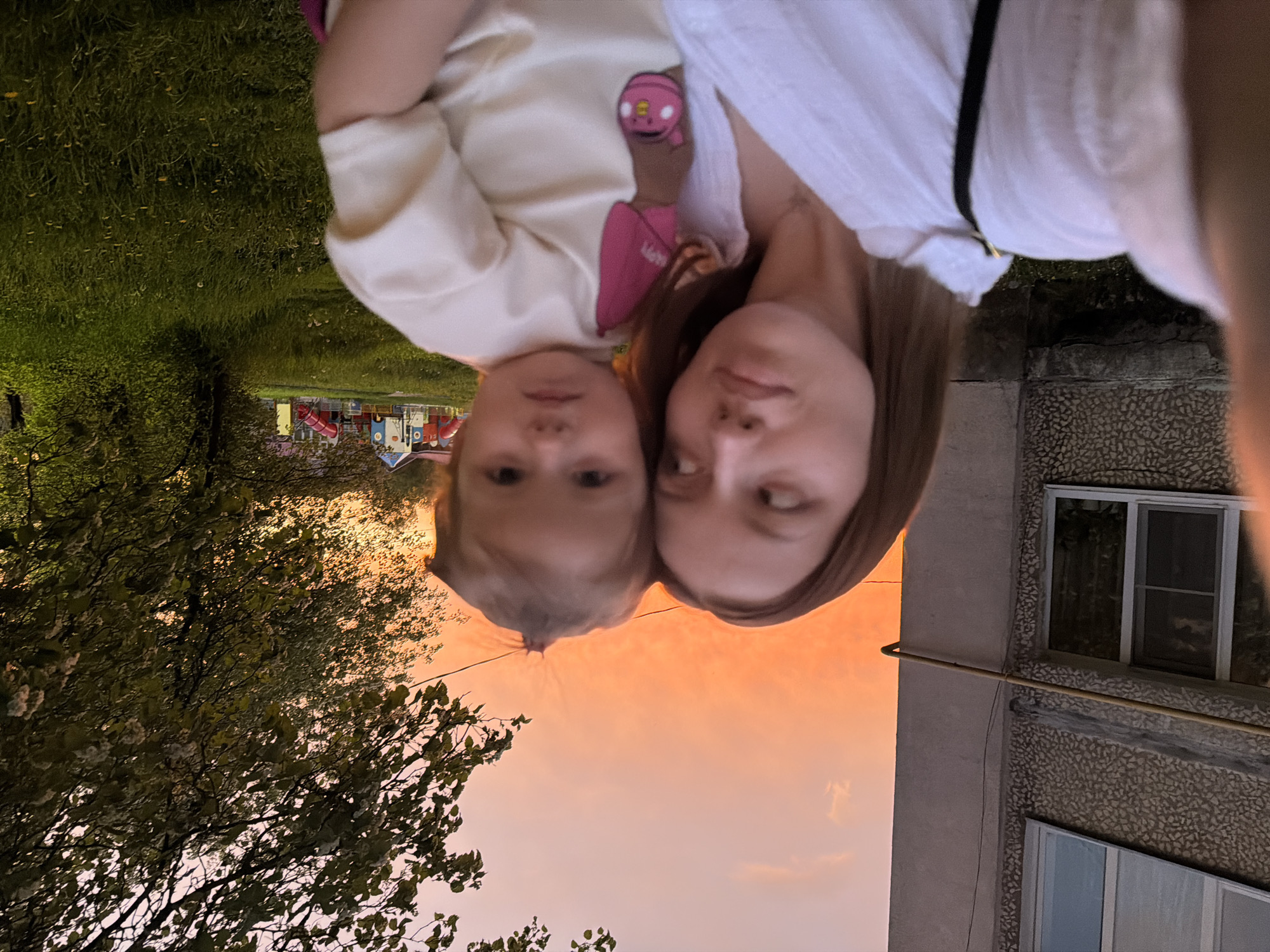 A woman and a child are visible in a selfie-style image. The background suggests an outdoor environment with a sunset sky, trees, grass, and a building. A playground is visible in the distance. The photograph appears to have been taken in Reshetnikovo, Russia.