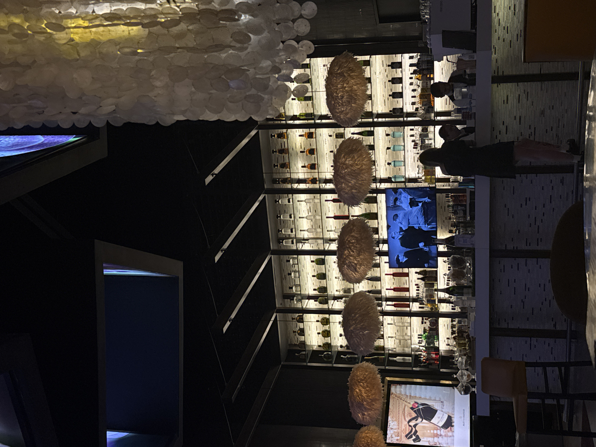 The image depicts the interior of a bar or restaurant in Playa del Carmen, Mexico. The main focal point is a back-lit bar shelving unit filled with various bottles of liquor and wine. Several decorative, possibly feathered, hanging lamps obscure the bottles. A television screen is mounted among the shelves, displaying images or video.
In the foreground, a couple is standing at the bar, possibly ordering drinks or conversing with the bartender. The bar is topped with glass and bar stools line one side.
Above, a ceiling decoration composed of numerous white, circular elements (possibly shell-like) partially obscures the view. The lighting is subdued, with the back-lit bar shelves providing the primary illumination. To the side, there is what appears to be an advertisement for Moët & Chandon champagne. A dark doorway or corridor is visible in the surrounding. The overall scene is indicative of a modern, upscale establishment.