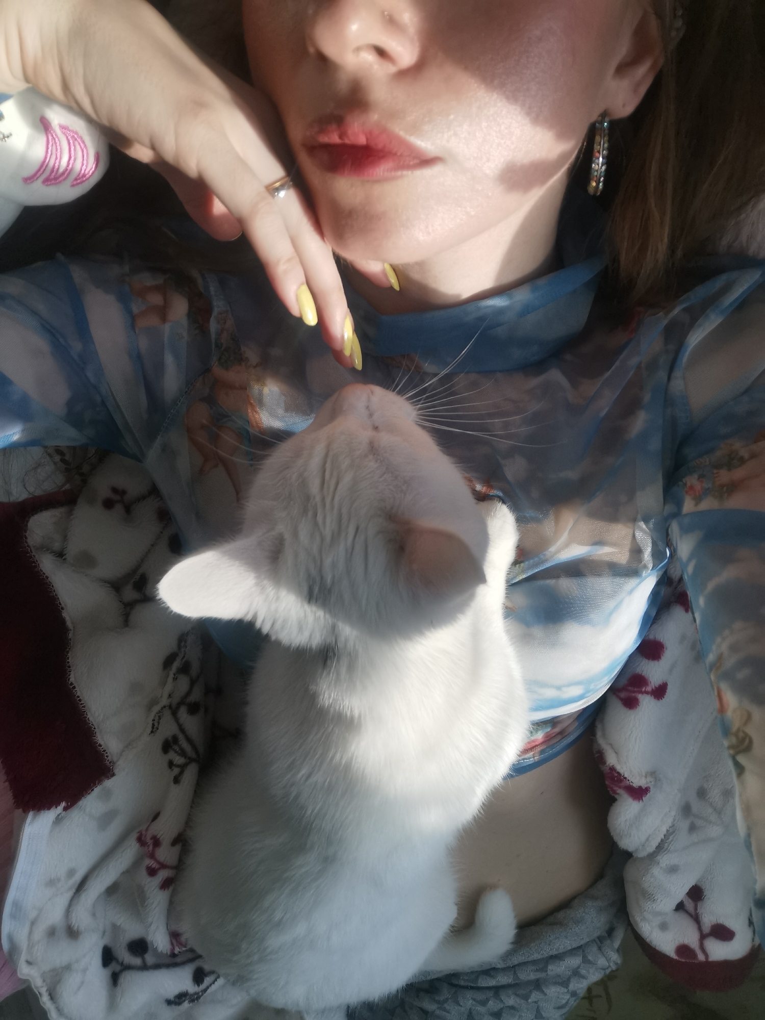 A woman is lying down indoors with a white cat resting on her chest. She has fair skin, red lips, and wears earrings. Her nails are painted yellow. She wears a blue, patterned top. The cat is white with a long tail. They are lying on a blanket or soft surface. The location is unknown.
