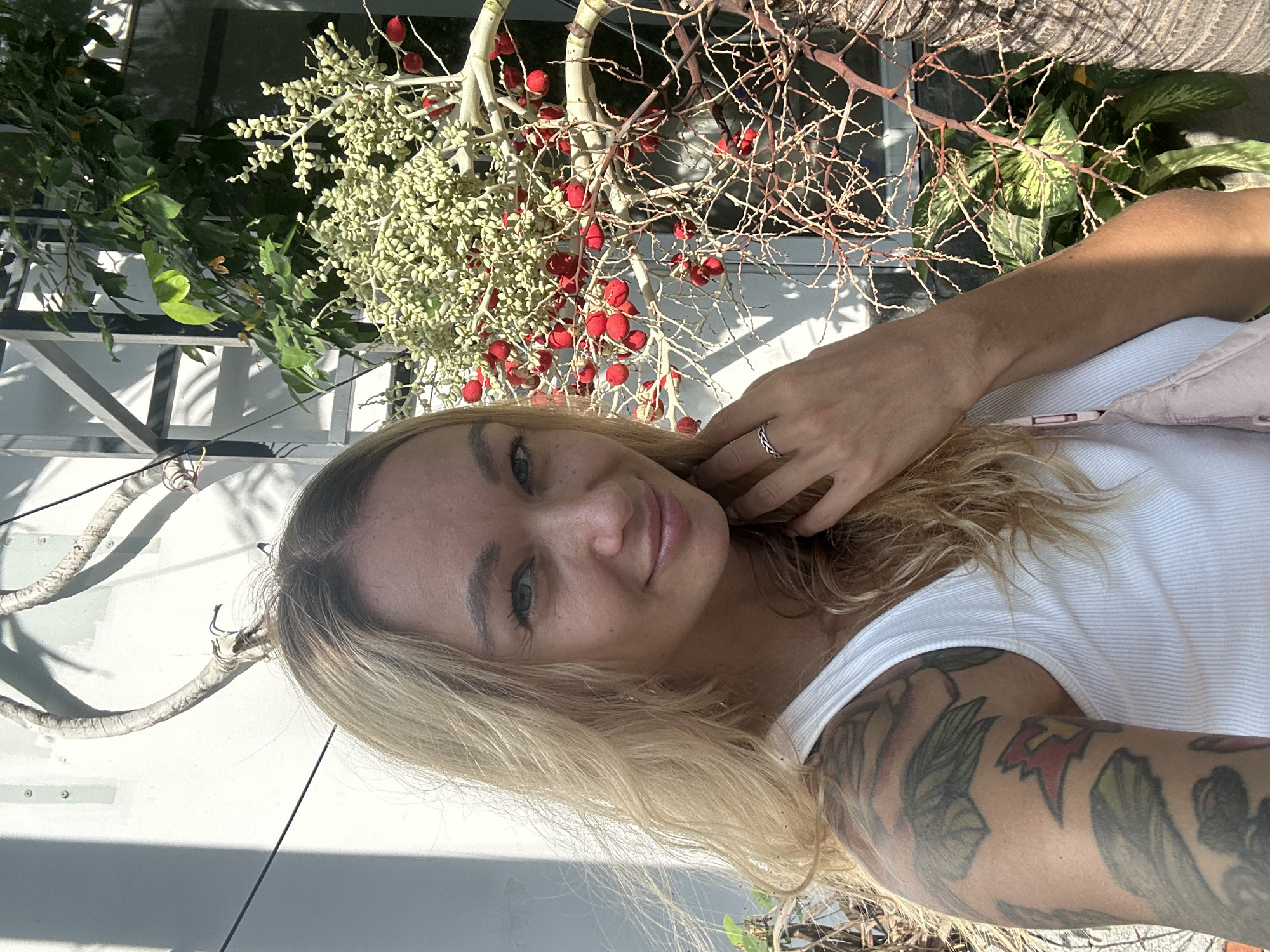 A blonde-haired female subject, with blue eyes and a slight smile, is positioned centrally, looking directly at the camera. Her hair is light blonde, wavy, and appears slightly disheveled. She wears a white ribbed tank top, and extensive, multi-colored tattoos adorn her left shoulder and upper arm, depicting foliage, possibly flowers, and a red and yellow emblem. A silver ring with a braided design is visible on the fourth finger of her left hand, which is lightly touching her neck/chin area. A light pink fanny pack or small crossbody bag strap is visible draped over her right shoulder.
The background consists of a light-colored wall on the left, illuminated by strong directional sunlight, casting distinct shadows of architectural elements (likely metal framing) and unseen structures. A white-wrapped conduit or cable system with exposed wires is visible high on the upper left wall. Dominating the right side of the frame is a branch from a palm-like tree, consistent with a Betel Nut Palm (Areca catechu), laden with multiple clusters of both unripe, pale green, ovoid fruits and fully ripened, vibrant red, spherical fruits. Behind this palm branch, darker green foliage is visible, along with a variegated plant with large green and cream-colored leaves (possibly a Dieffenbachia) at the lower right. The scene is outdoors, brightly lit, indicating a tropical or subtropical environment, consistent with the city of Mũi Né, Vietnam.