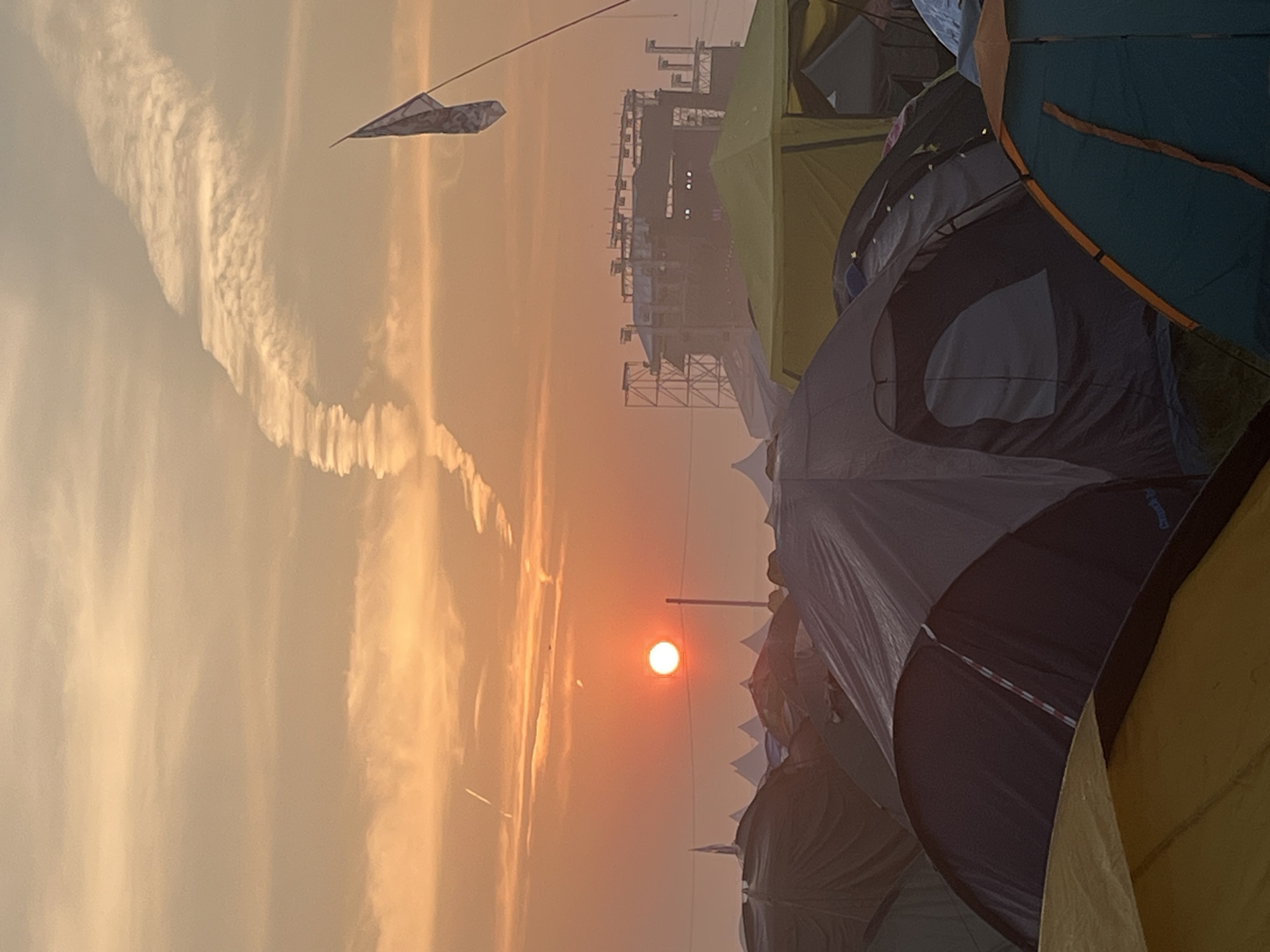 The image captures a sunset scene over a campsite, with numerous tents in the foreground and a large stage structure in the background. The sky is a vibrant orange and yellow, with wispy clouds and a prominent contrail. The sun, a bright orange orb, is setting on the left side of the frame. Several tents of various colors and sizes are pitched closely together, suggesting a festival or event. A flag is visible on a pole to the right of the stage structure. The stage appears to be made of metal scaffolding and has lights installed. The overall impression is one of a large outdoor gathering at dusk. The specific location in the city of Surakovo, Russia, cannot be confirmed from the visual information provided in the image itself.