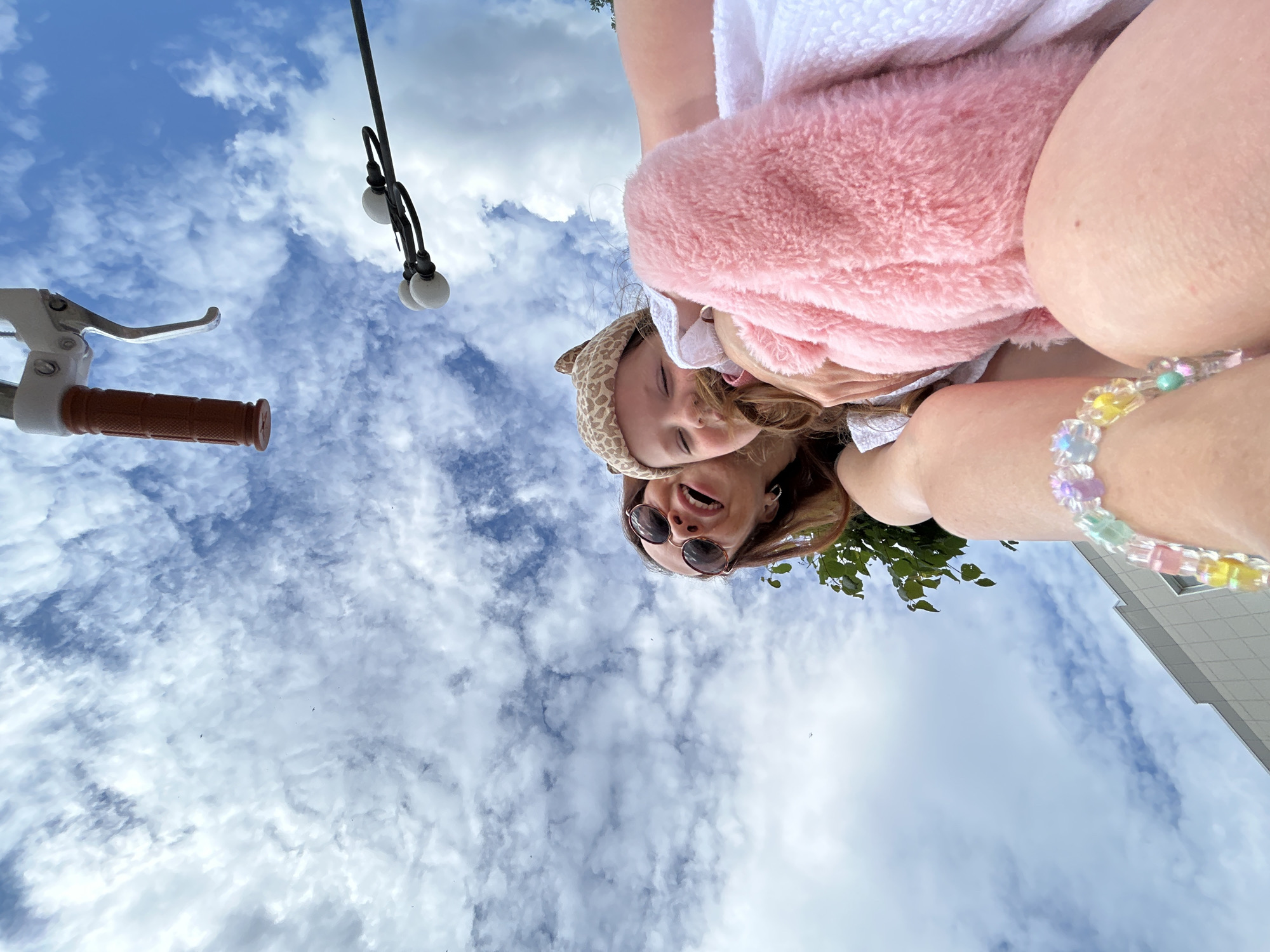 In an upward-facing, wide-angle shot taken from a low perspective, a woman and a young child are partially visible against a backdrop of a cloudy blue sky. The woman, on the left, is wearing sunglasses and has her mouth open in what appears to be laughter. She has long, brown hair. The child, positioned to the right and slightly in front of the woman, is wearing a leopard-print hat and is nestled in a pink fuzzy blanket. The child's eyes are closed, and their hair is being touched by the woman. A part of a bicycle handlebar with a brown grip is visible at the top of the frame, suggesting the scene might be from a bicycle ride. In the upper right corner, a street lamp with three white globes is visible. At the bottom left, the corner of a building with a grid-like facade is visible. In the lower right, a person's leg is visible, adorned with a colorful beaded bracelet. The image was captured on April 29, 2023. The location is specified as Chaikovsky, Russia.