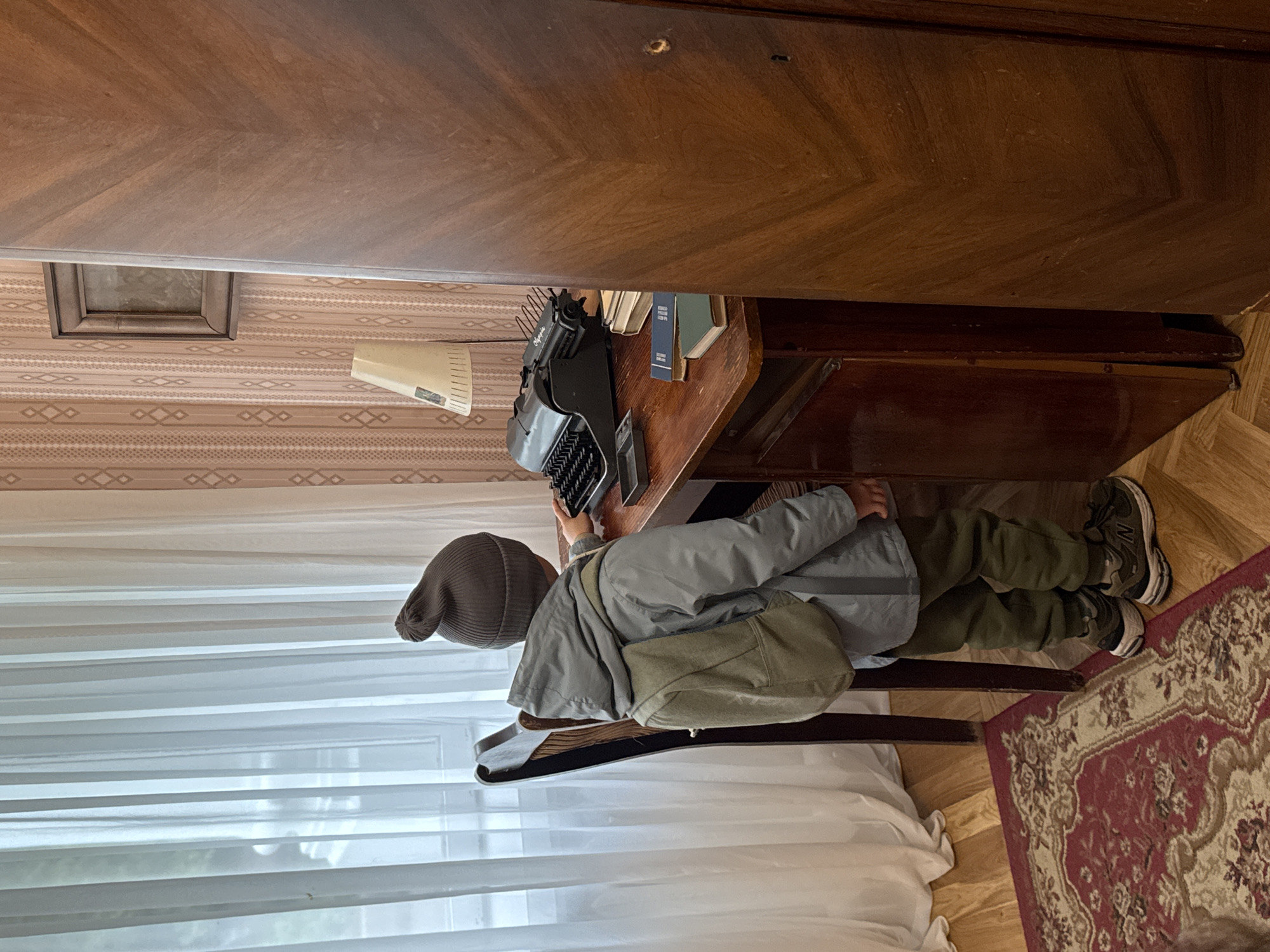 A person wearing a gray hat, a gray jacket, and olive green pants sits in a chair in front of a desk with a typewriter on it. The desk is made of dark wood and has books on it. The room has patterned wallpaper, a wooden floor, and a rug with a floral design. A window with white curtains is to the left of the person. The photograph was taken at the Andrei Tarkovsky Memorial House in Vnukovo, Russia.