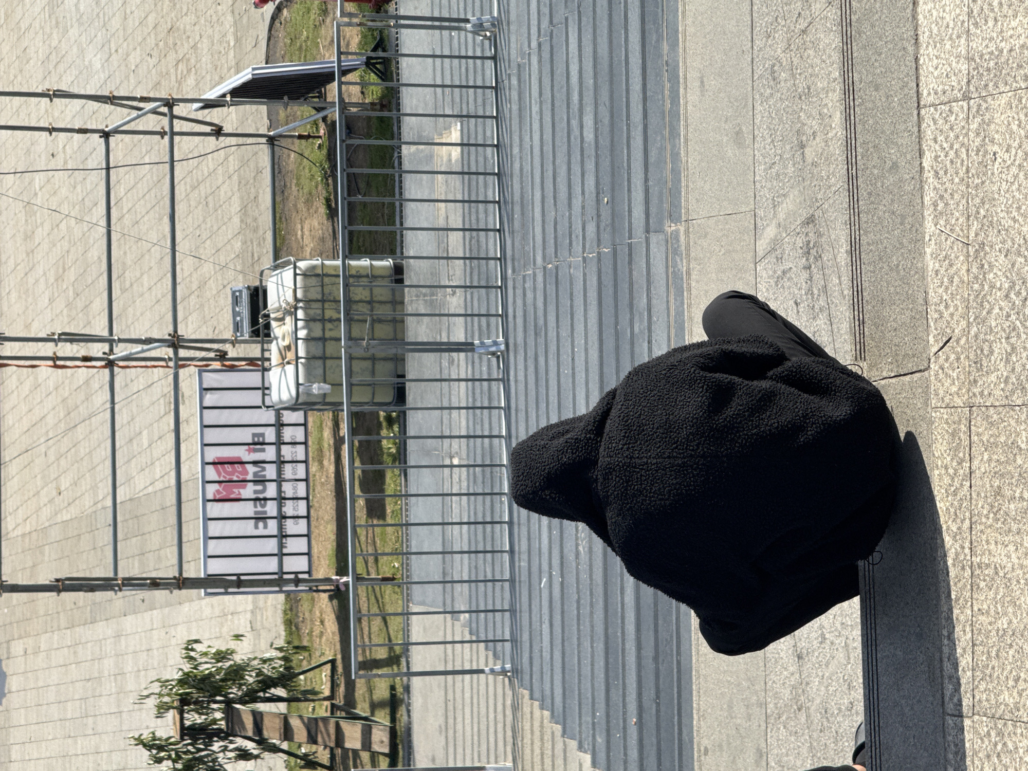 A person wearing a black hooded jacket is sitting on gray, stepped stairs, facing away from the camera. The background shows an outdoor urban scene with a tiled ground, a metal fence, and scaffolding. A white banner with red lettering is visible behind the fence, and a large white water tank is positioned near the scaffolding. To the left, a wooden ramp with a small tree is also visible. The overall lighting suggests it is daytime with strong shadows cast by the sun. The location is not identifiable as Da Lat, Vietnam.