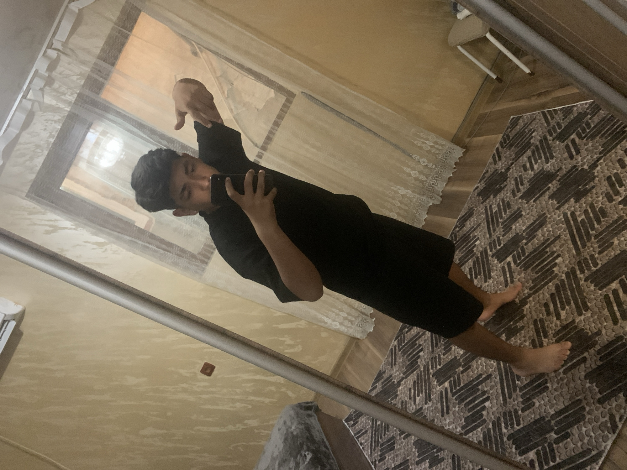 A reflection of a person taking a selfie is visible in a mirror. The person is wearing a black top and shorts. They are holding a smartphone and pointing with their other hand. The background includes a curtained window, a beige wall, a patterned rug, a small chair, and part of a room interior. The location is potentially in Stara Zagora, Bulgaria, but there is no definitive evidence to confirm this.