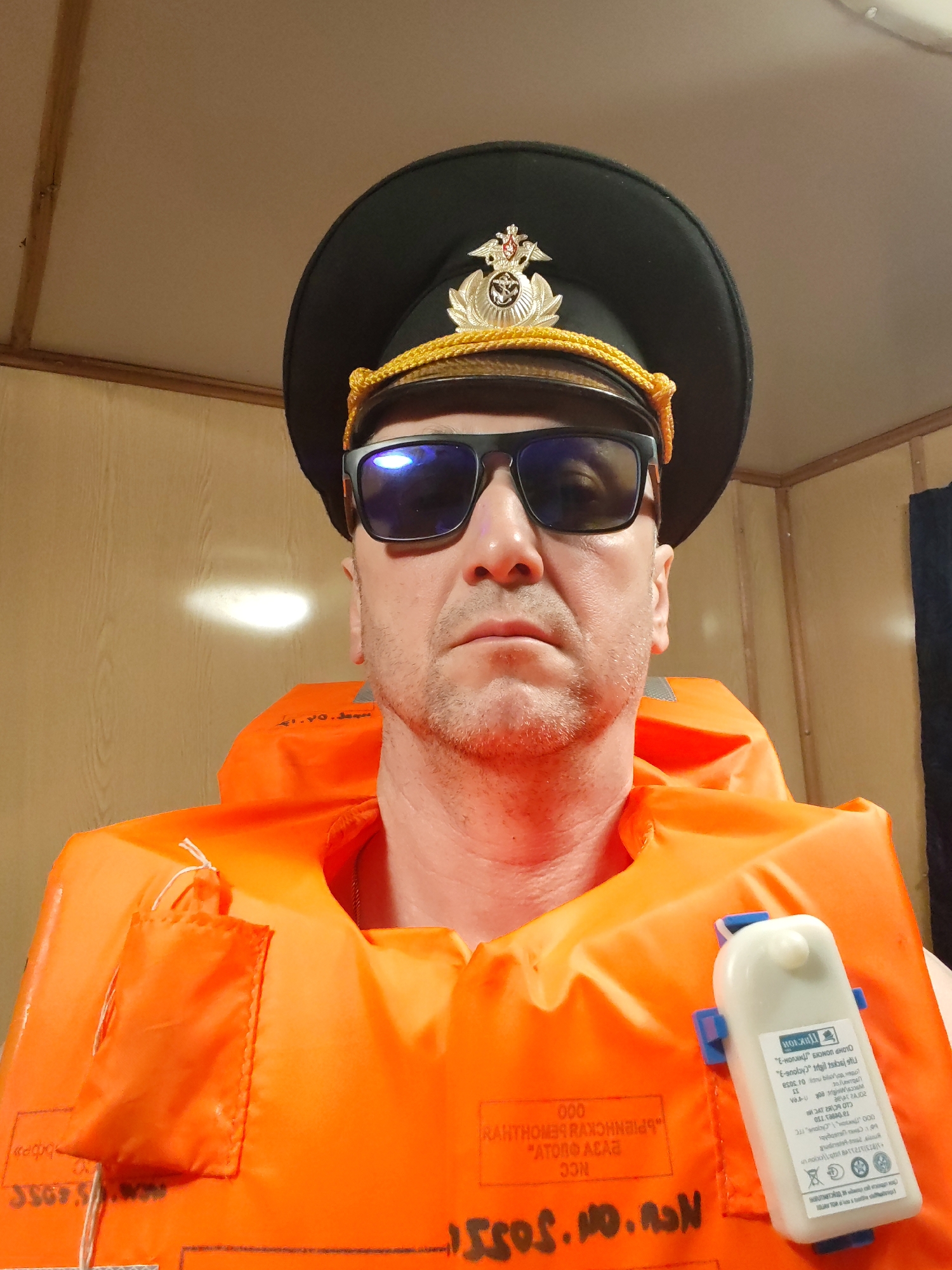 A man is wearing a black hat with a gold-colored emblem and trim, black sunglasses, and an orange life vest. The vest has white writing and a small white device attached to it. The background is plain with vertical wood paneling. The exact location and city are not discernible from the image.