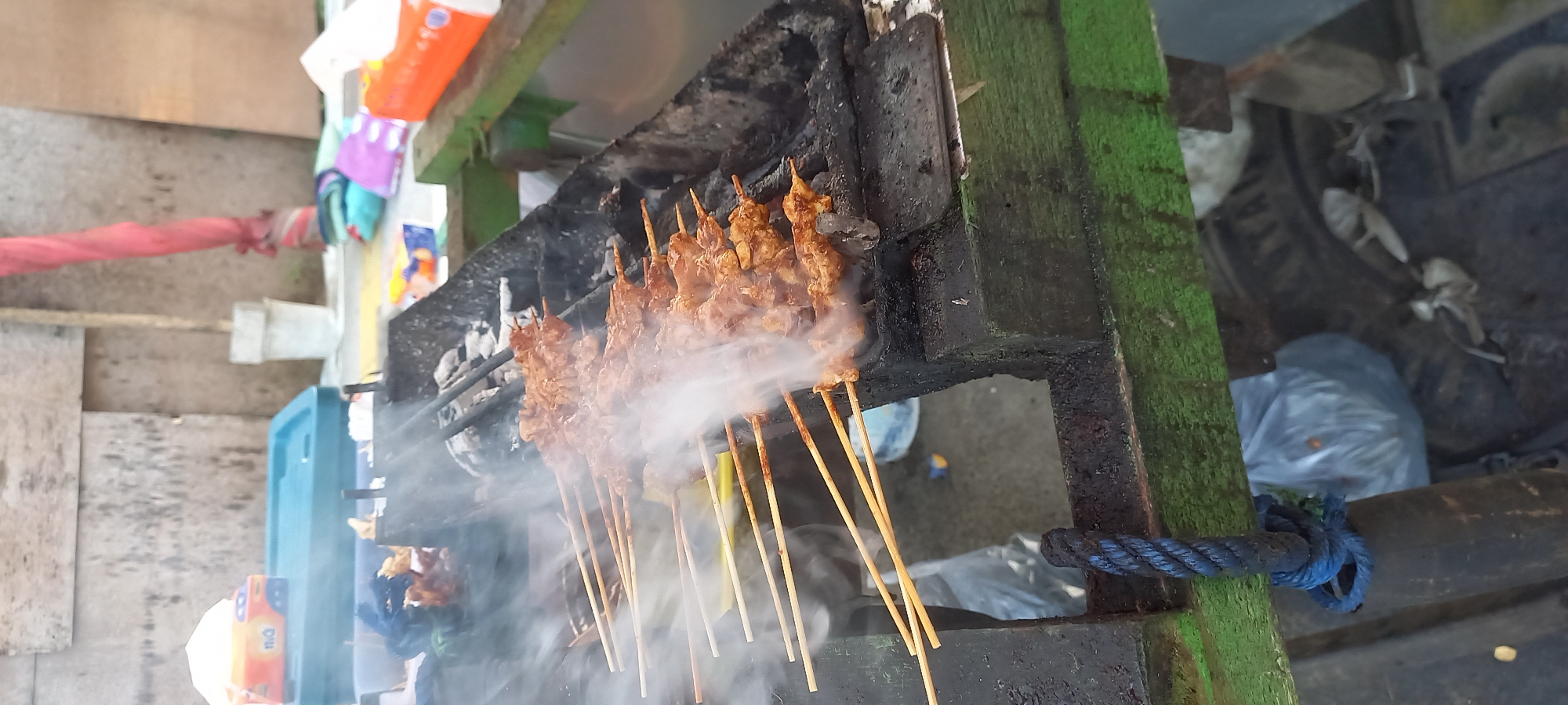 A charcoal grill is positioned in the foreground, laden with skewers of satay, emitting a thick plume of white smoke. The grill is set upon a green wooden structure, part of a mobile food cart. To the left, a blue rope is tied to the cart. In the background, a weathered wall is visible, with a red cloth draped over a pole and various colorful items scattered on a table. Tissue boxes, one orange and one with a lighter blue hue, are also present on the table, along with a turquoise plastic container. The setting appears to be an outdoor market or street stall in Panji, Indonesia.