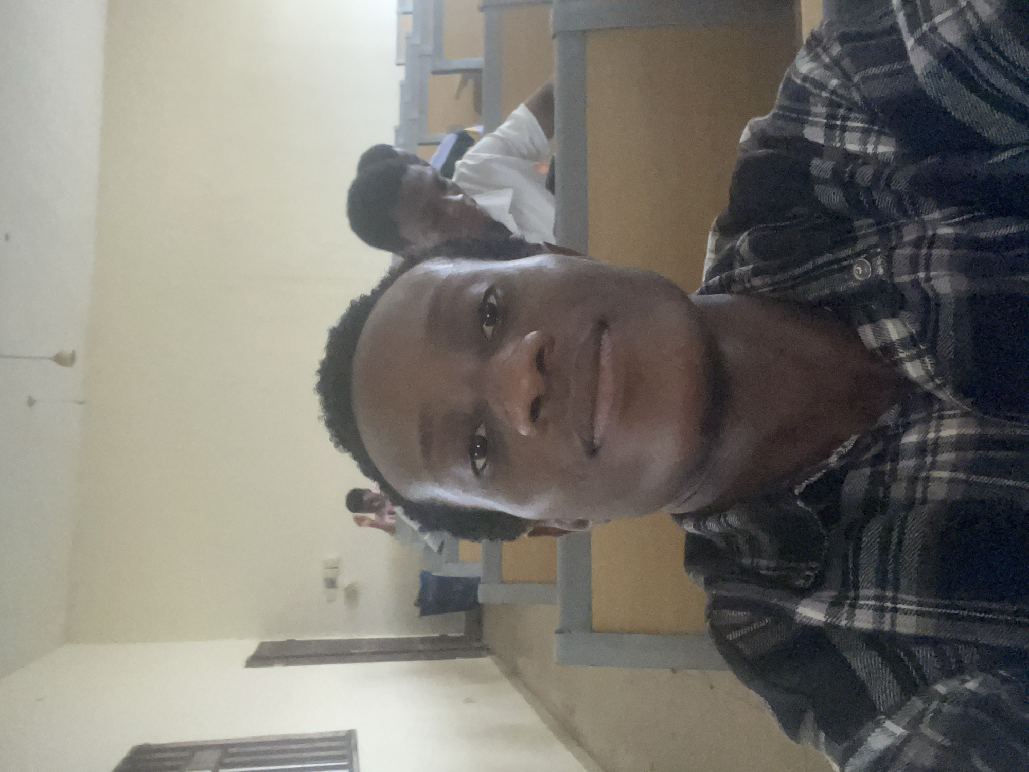 In a classroom setting in Ihiagwa, Nigeria, a young Black man is captured in a selfie. He is positioned in the foreground, occupying the majority of the frame, with a portion of his head and shoulders visible. He is wearing a dark and light plaid shirt. Behind him, two other individuals are seated at desks. The room appears to be a lecture hall with rows of light brown and gray desks. The walls are painted a pale cream color, and a window with dark bars is visible on the left side of the image. A dark blue trash can is also present in the background. The lighting suggests an indoor environment with overhead illumination.