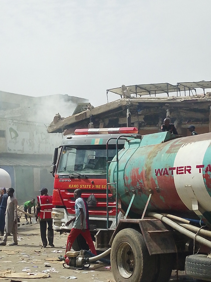 The scene in Fagge C, Nigeria, depicts an ongoing emergency response. In the foreground, a red HOWO truck marked "KANO STATE FIRE SERVICE" is positioned next to a large, weathered water tanker truck, painted teal, red, and white, with "WATER TANKER" stenciled in red. Hoses and a portable gasoline-powered pump are visible on the debris-strewn ground between the vehicles. Several individuals are present: one person in a red safety vest stands near the fire truck, another in a light blue shirt and red trousers walks past the pump, and two individuals are observed on top of the water tanker. In the background, to the left, smoke emanates from a partially collapsed multi-story structure. The roof of a building to the right shows exposed support beams and what appears to be a structure for solar panels. The environment is dusty, with numerous papers and small pieces of refuse scattered across the ground. The sky is bright and hazy.