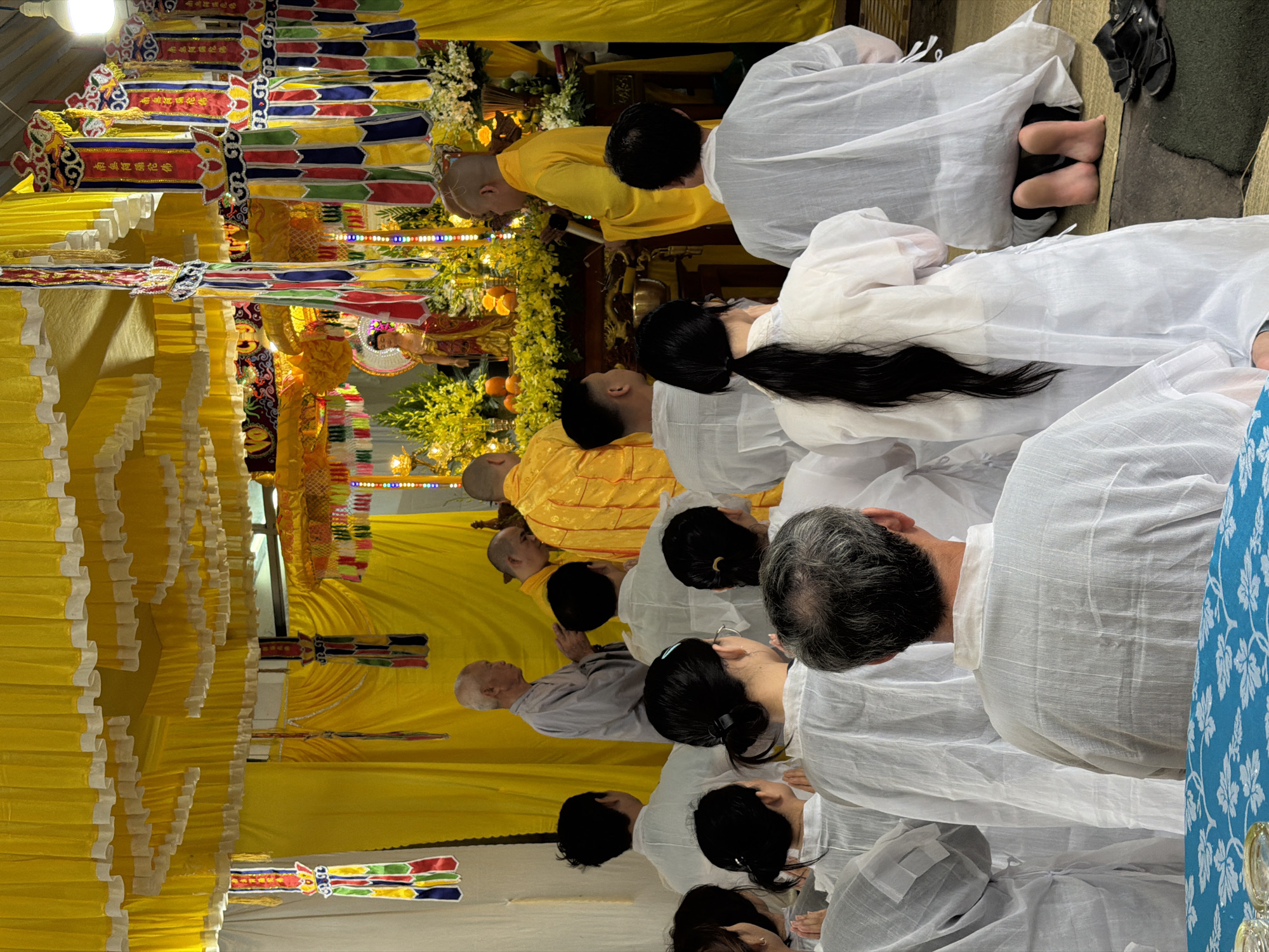The image depicts a group of people, mostly dressed in white, kneeling in a line, possibly at a religious ceremony or funeral. There is a prominent altar decorated with yellow drapery, flowers, and religious symbols. Some figures are wearing yellow robes, suggesting they are monks or religious officials. The setting is likely indoors, possibly a temple or a home adapted for a religious service.
It's difficult to confirm the location specifically as Đà Nẵng, Vietnam without additional context, but the decorations, robes, and kneeling arrangement are consistent with funeral or religious practices common in Vietnamese culture.