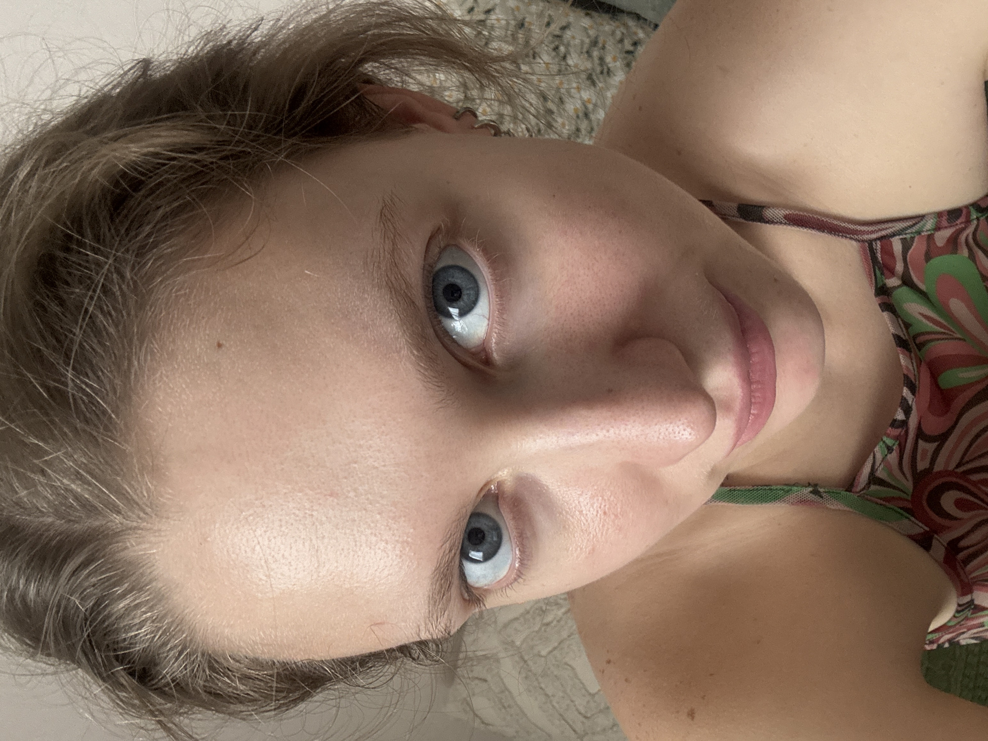 This is a close-up, high-angle, selfie-style photograph of a young Caucasian woman's face, taken indoors. The subject has fair skin with visible pores and some freckles. Her eyes are large and blue with a direct gaze towards the camera. Her hair is light brown, appearing somewhat tousled, and is pulled back from her face. She is wearing a patterned tank top with green, pink, and white swirling designs. The background is blurred and indistinct, with a textured surface that appears to be upholstery or fabric. There is no indication of the specific location being Izhevsk, Russia.