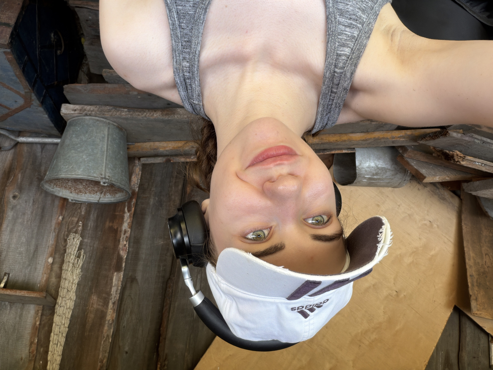 A young woman, wearing a grey tank top, a white Adidas baseball cap, and black headphones, takes a selfie. She has light skin, green eyes, and dark brown hair tied back in a braid. The background appears to be an outdoor shed or workshop, with wooden walls and shelves. On the shelves are various objects, including a metal bucket, a piece of netting, and some scrap wood. The lighting suggests it is daytime, with sunlight illuminating the scene. The woman is smiling faintly, and her gaze is directed towards the camera. This scene is not specifically located in Zavodoukovsk, Russia.