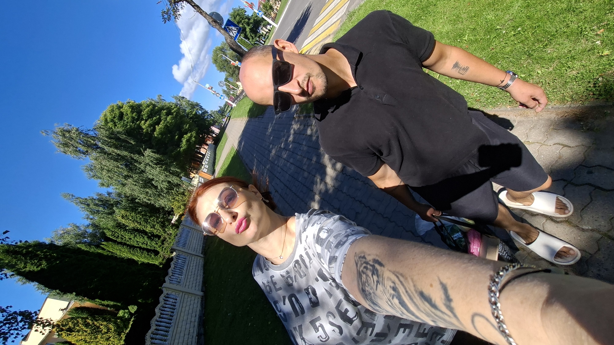 Two individuals, a woman with red hair and sunglasses, and a man with sunglasses, are taking a selfie outdoors on a paved walkway bordered by grass. The woman is wearing a grey t-shirt with a pattern and the man is wearing a black polo shirt and black shorts. They are both wearing sandals. The background includes trees, a white fence, and a road with a crosswalk. The sky is blue with some clouds. There are shopping bags near the man's feet.