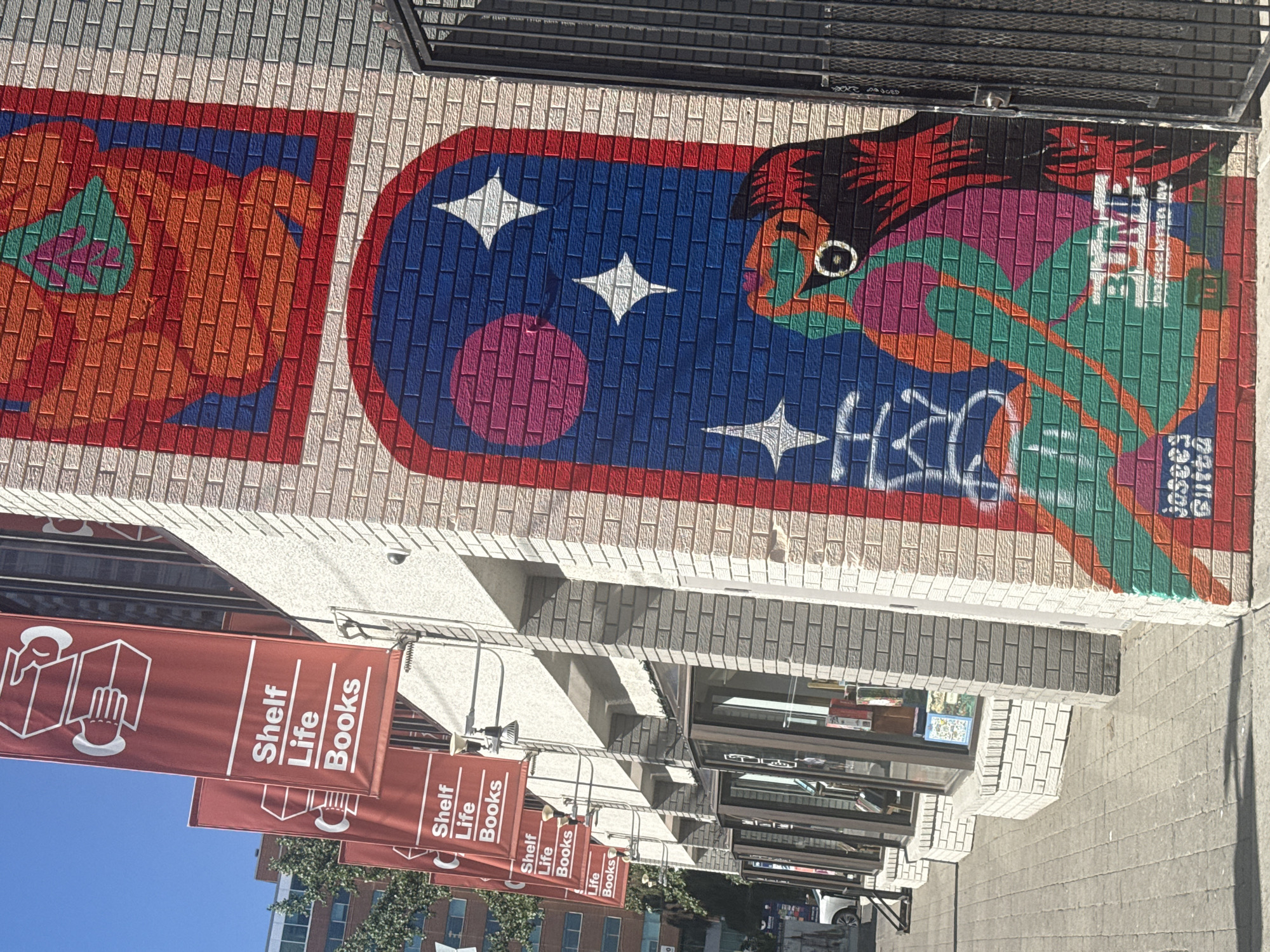 This image captures the exterior of "Shelf Life Books," a bookstore located in Calgary, Canada. The building features a light-colored brick facade adorned with vibrant murals. Three red banners hang vertically along the side of the building, each displaying the store's name, "Shelf Life Books," in white lettering and an illustration of hands holding books.
The murals are colorful and abstract. The upper mural, on a corner of the building, features a red border enclosing a geometric design in shades of orange, red, and green. Below this, a larger mural depicts an arched window opening onto a starry night sky. The sky is painted in dark blue with pink and white stars, and a large pink circle resembling a moon or planet. Within the archway, a stylized bird-like creature with large, round eyes and feathered wings is painted in a spectrum of colors including teal, pink, orange, and red. The artist's signature, "Josees Guttz," is visible in the lower part of this mural, along with the tag "3UMB" and "EASTER AV."
In front of the building, a paved sidewalk runs along the storefront. Several large windows display books and merchandise inside the store. A black metal gate stands to the right of the mural. The sky is clear and blue, indicating a sunny day. The overall scene conveys a lively and artistic atmosphere for the bookstore.