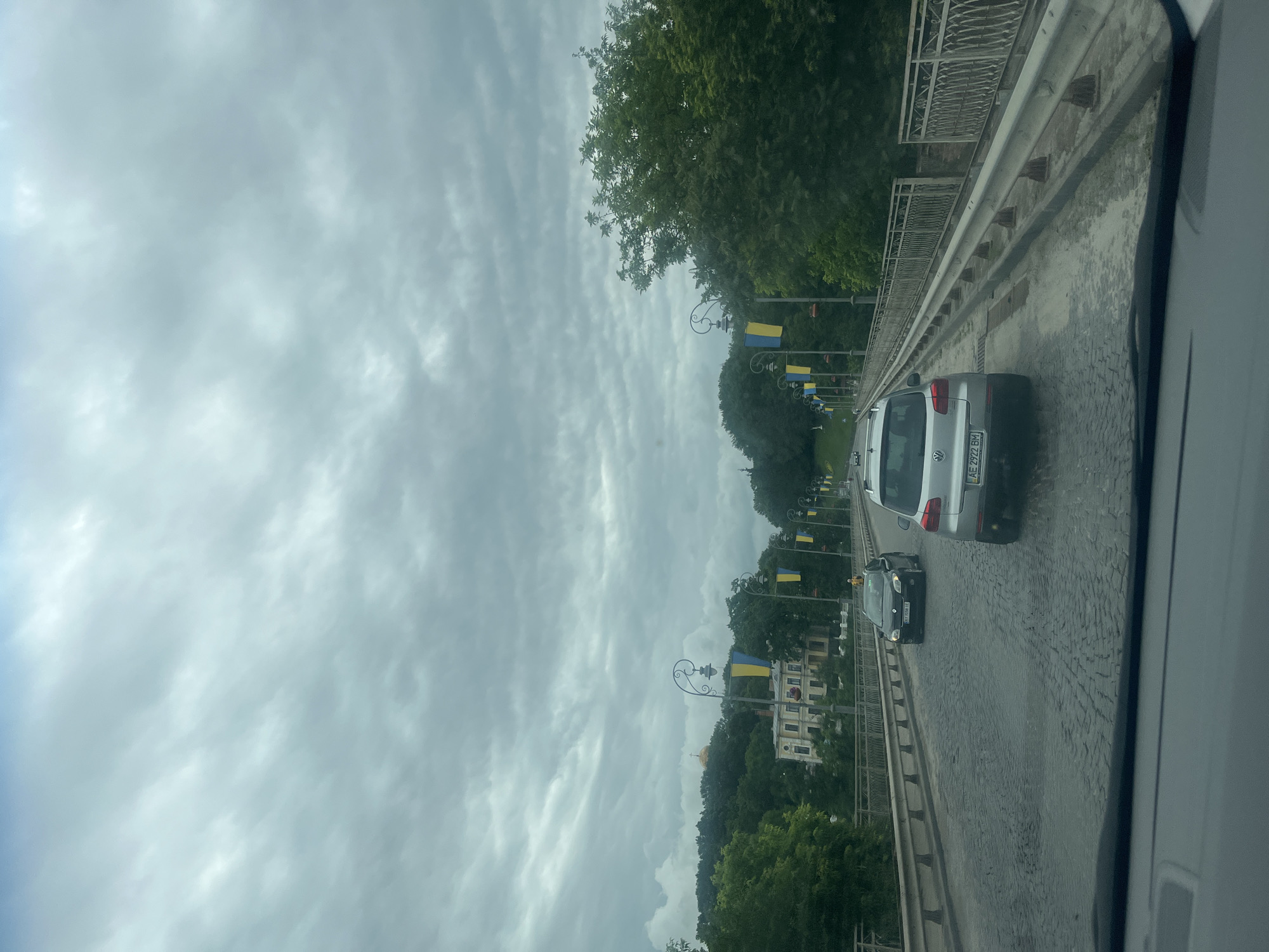 The image shows a view from a vehicle driving on a cobblestone bridge in Kamianets-Podilskyi, Ukraine. Ukrainian flags are displayed on lampposts lining the bridge, and lush green trees and a building with a golden dome are visible in the background. Two cars are driving on the bridge, and the sky is overcast.